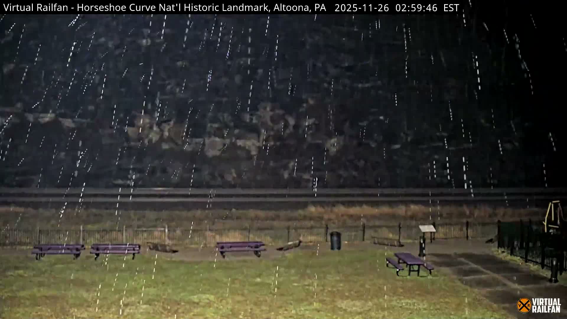 The image depicts an outdoor park area at night with several purple benches, a picnic table, and a fence, with train tracks and a dark wooded hillside in the background, under conditions of heavy falling rain or snow.