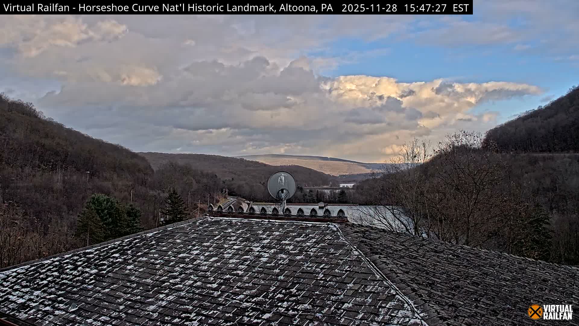 A cold, partly cloudy day reveals a snowy shingled rooftop with a satellite dish in the foreground, overlooking a vast valley filled with bare forested hills and a distant body of water, under a sky of scattered white and blue clouds.