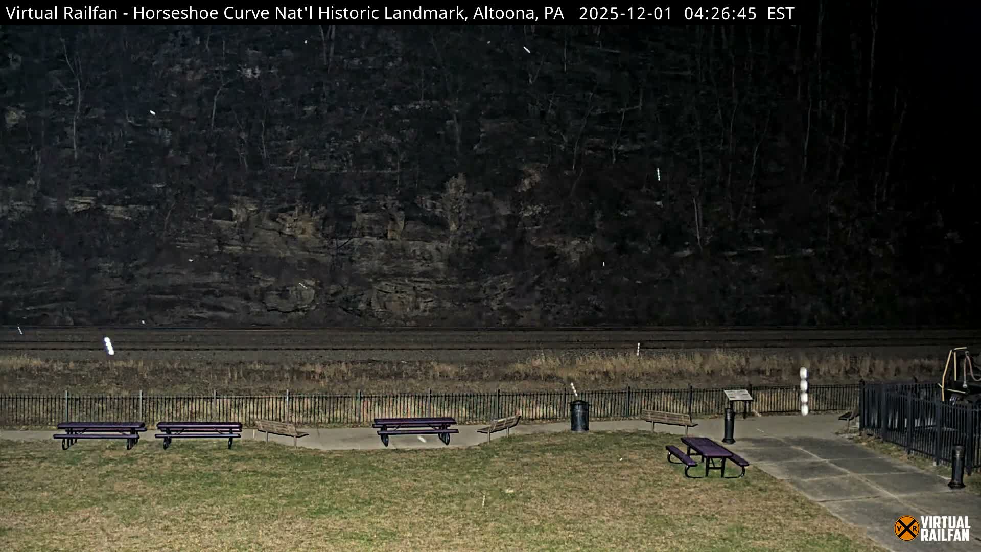 A dark, outdoor scene at Horseshoe Curve displays picnic tables and benches on grassy ground in the foreground, with railroad tracks and a rocky, tree-covered hillside in the background, all illuminated by minimal light under what appears to be a light snowfall.