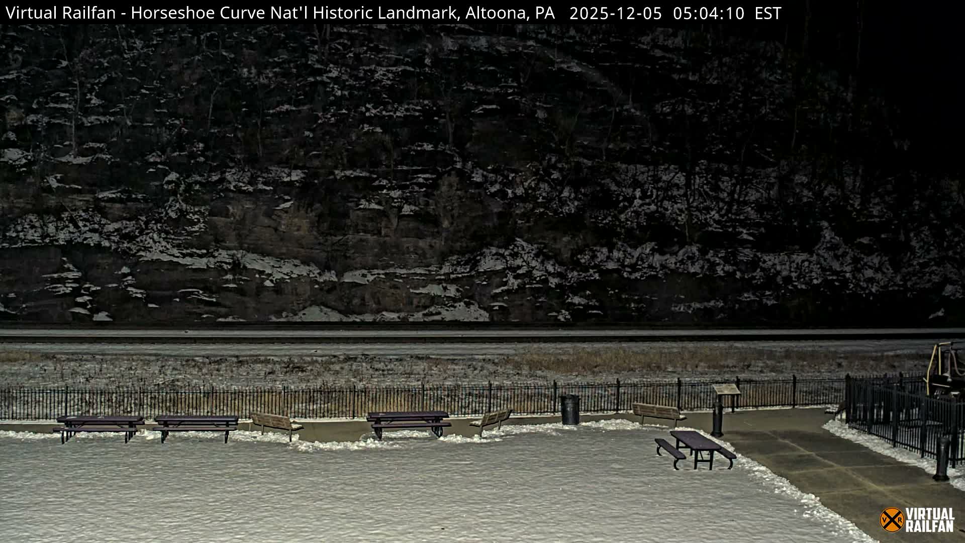 A snow-dusted outdoor railway viewing area, featuring benches and train tracks, lies at the base of a dark, snow-covered hillside during a cold night or early morning.