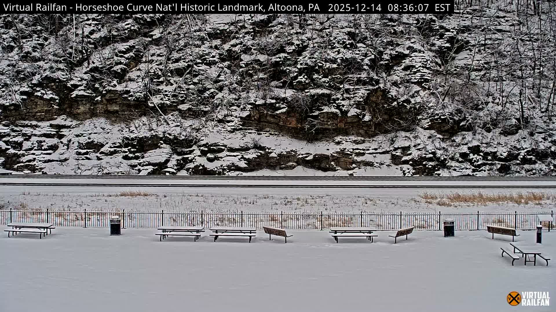 A snow-dusted outdoor railway viewing area, featuring benches and train tracks, lies at the base of a dark, snow-covered hillside during a cold night or early morning.