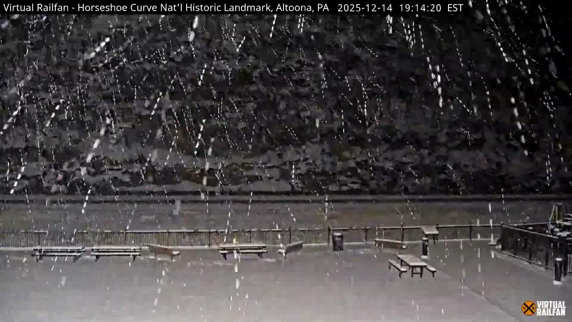 A snow-dusted outdoor railway viewing area, featuring benches and train tracks, lies at the base of a dark, snow-covered hillside during a cold night or early morning.