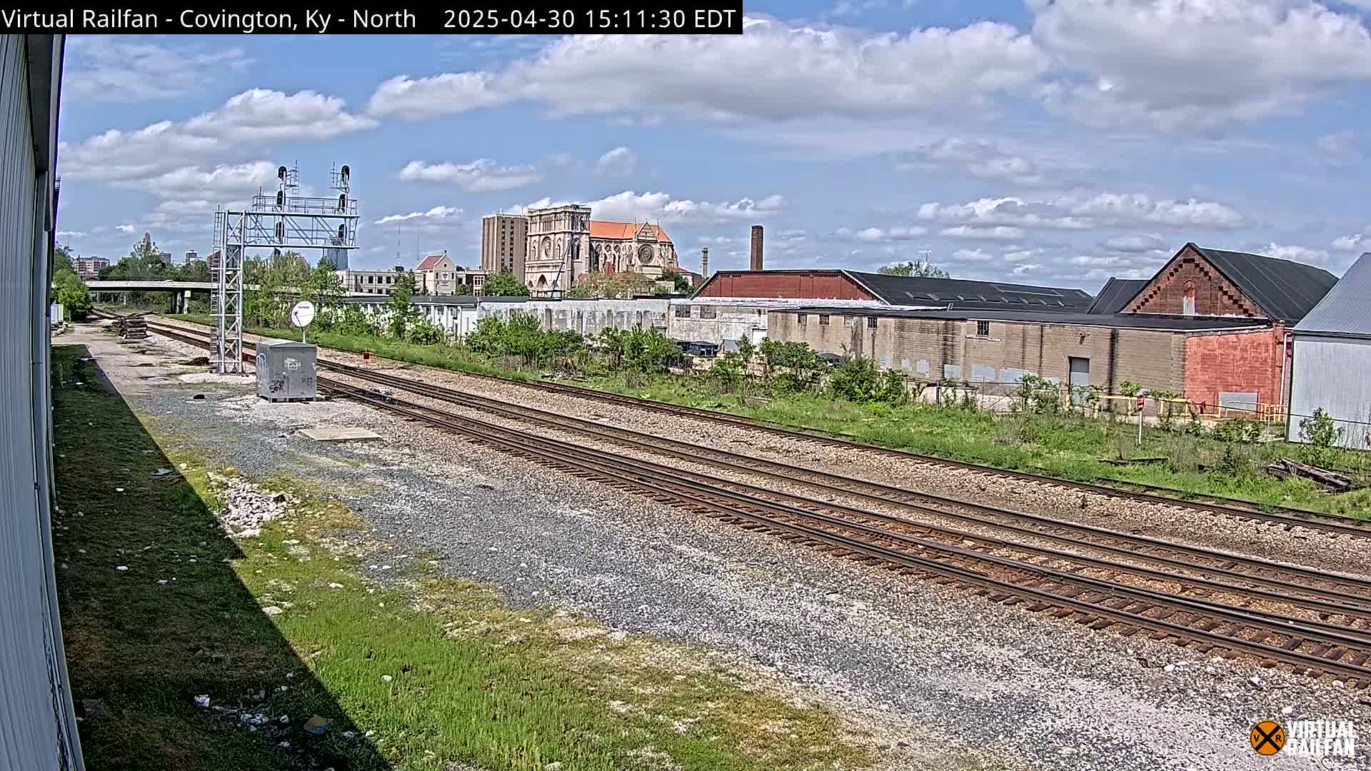 A partly cloudy day reveals railroad tracks stretching towards a cityscape featuring industrial buildings and a large church, all surrounded by patchy green vegetation.