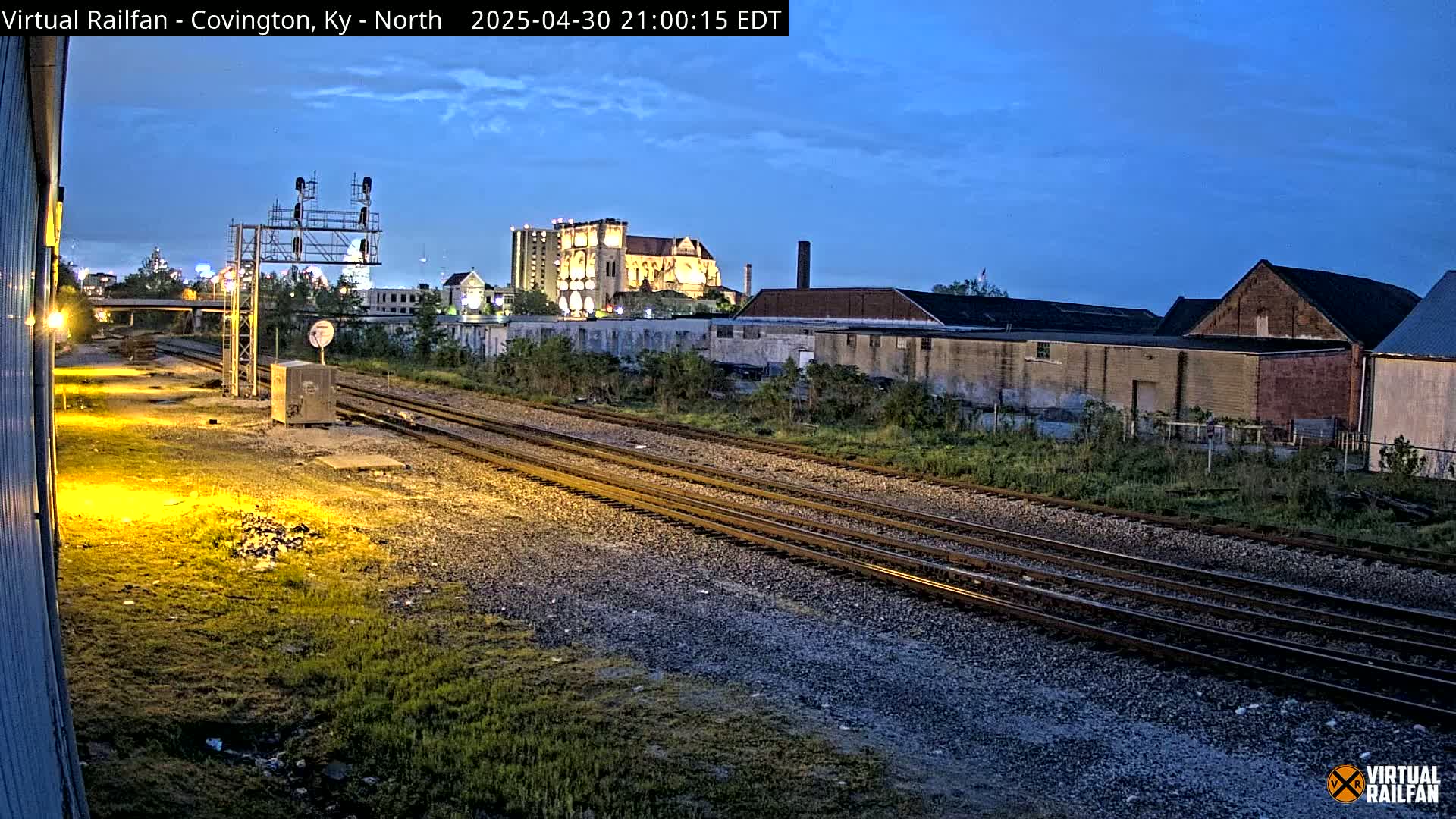 Under a twilight sky, three sets of train tracks run through a flat, grassy area toward industrial buildings and a large illuminated building in the distance.