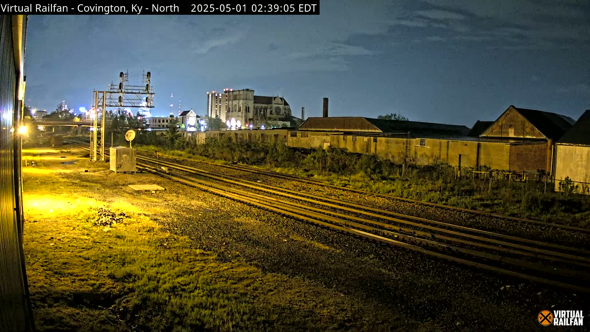 Under a dark, night sky, several sets of train tracks curve through a mostly empty, grassy lot toward some industrial buildings and a cathedral-like structure in the distance.