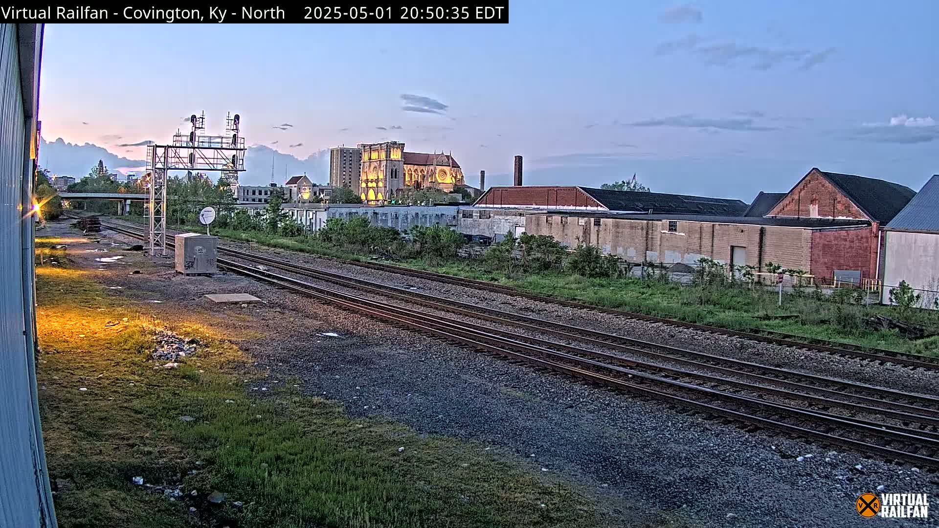Railroad tracks run through a grassy area towards a city skyline at dusk, featuring a large cathedral and industrial buildings.