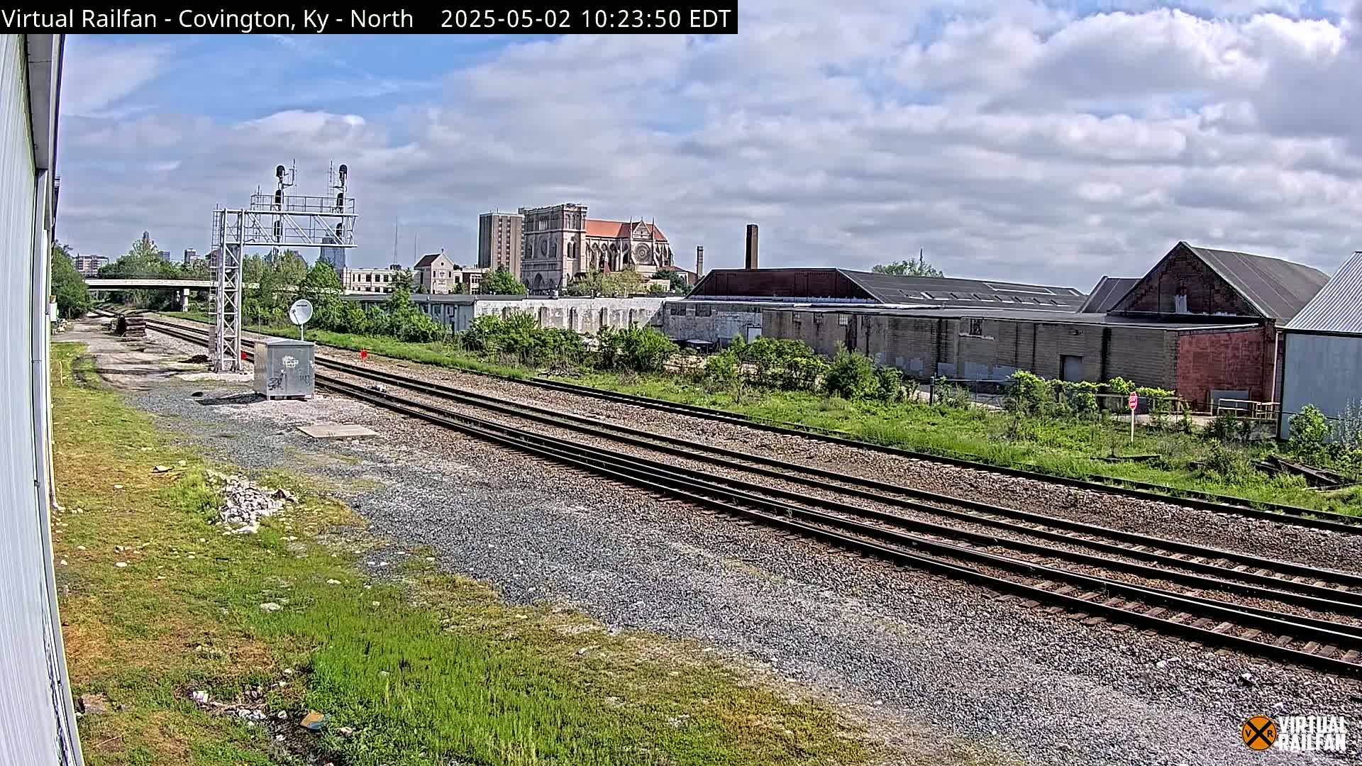 A view of railroad tracks on a partly cloudy day, with industrial buildings and a large church visible in the background.