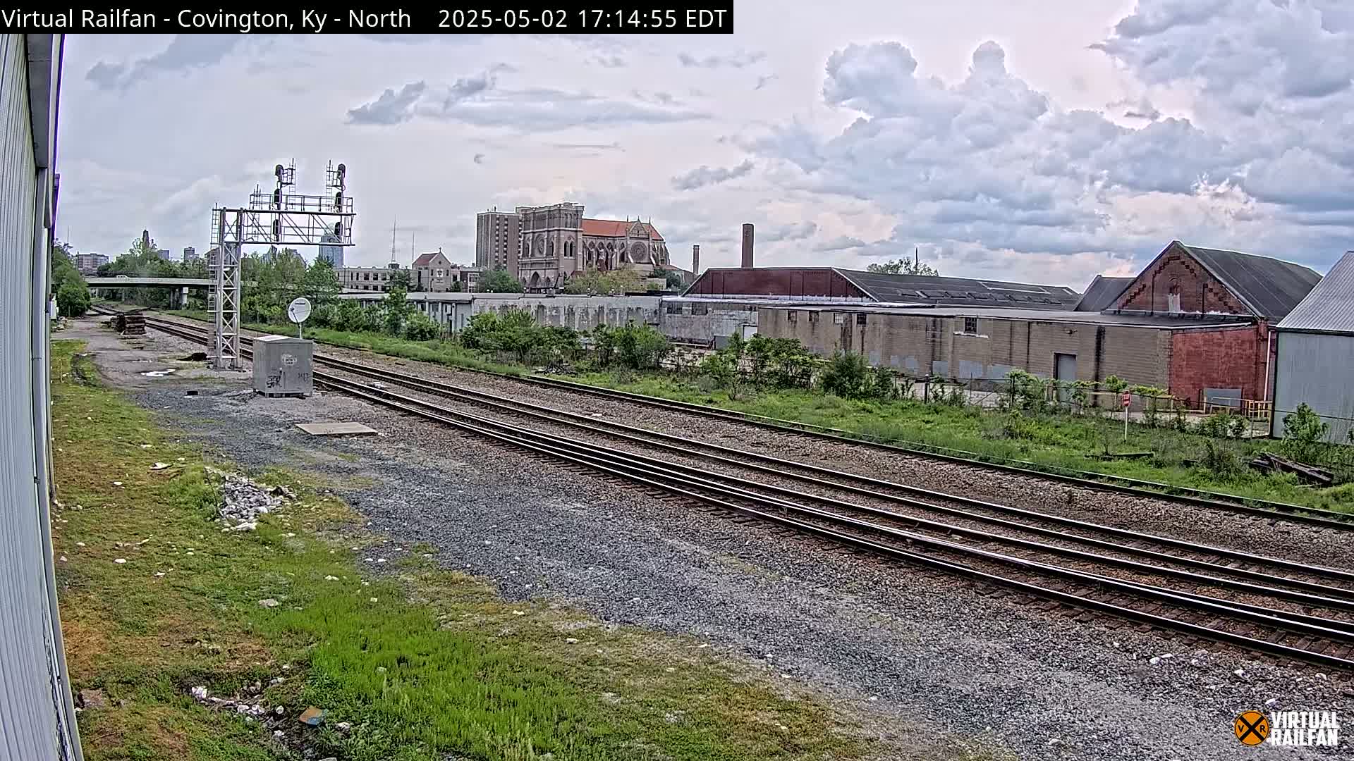 A view of railroad tracks under a partly cloudy sky, with industrial buildings and a church visible in the background.
