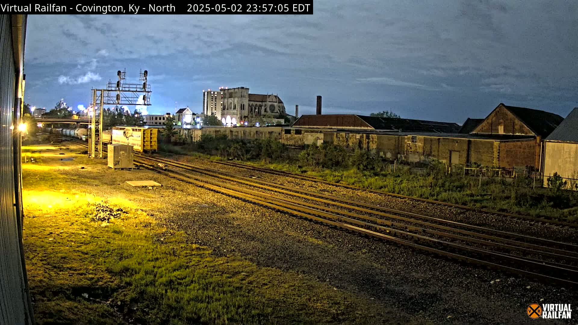 Under a twilight sky, a nighttime view shows a freight train partially visible on multiple sets of railroad tracks near several industrial buildings.