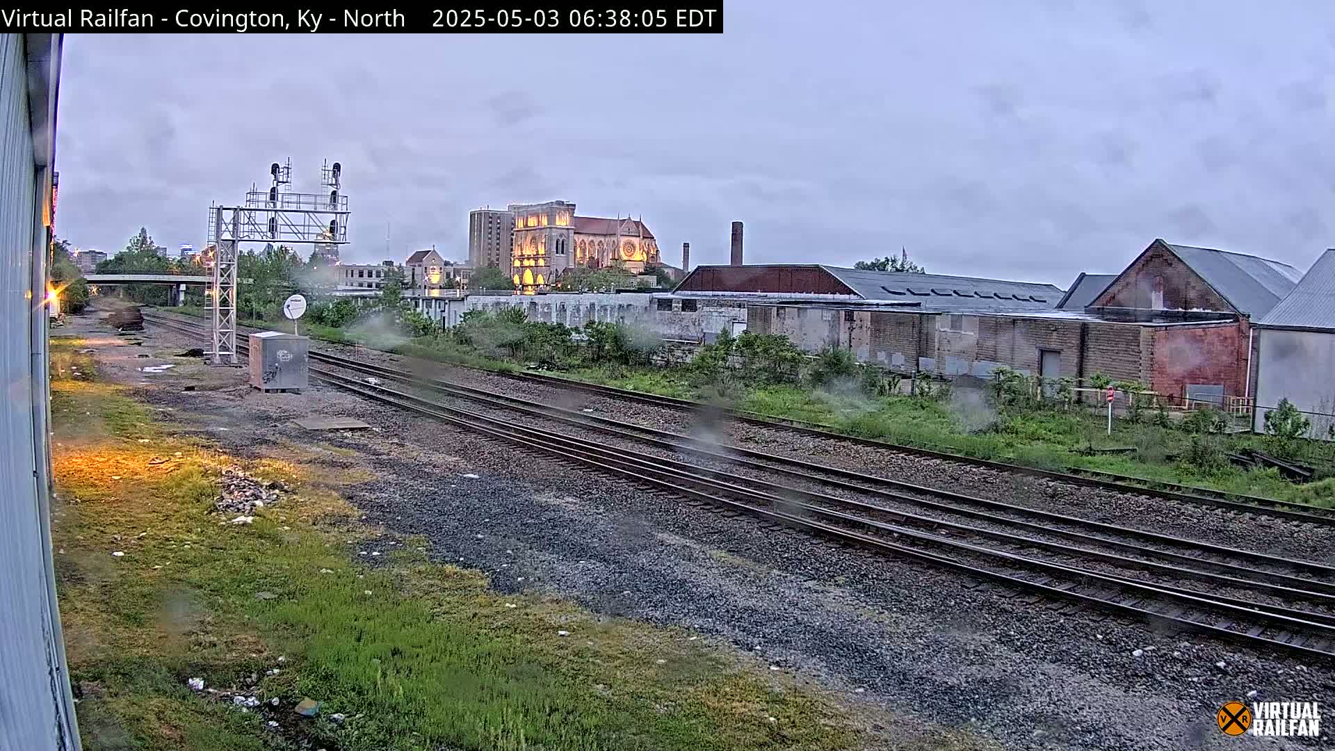Multiple sets of train tracks run through an overgrown area, past industrial buildings and a large cathedral under an overcast sky.