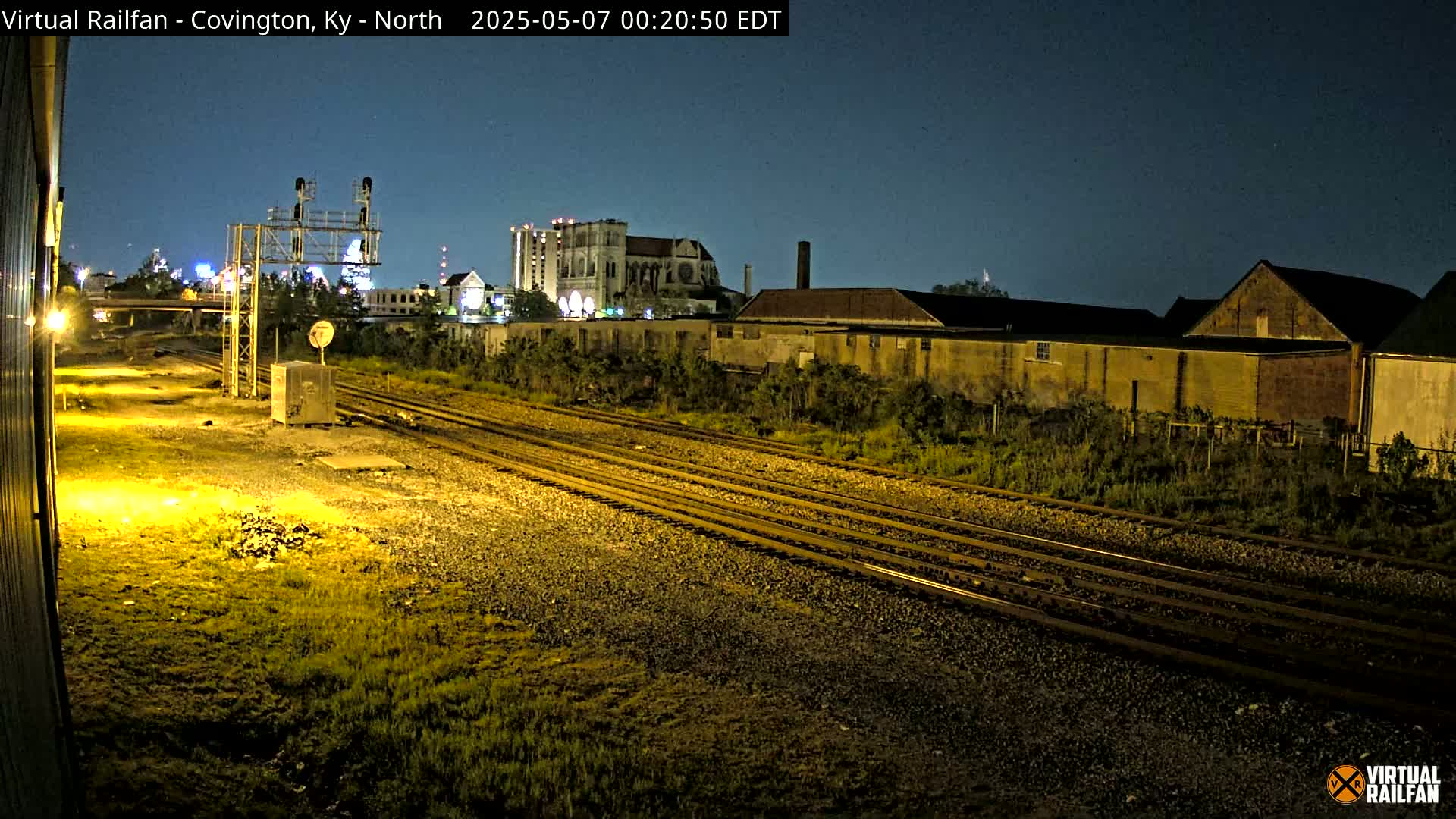 Under a dark night sky, multiple sets of train tracks extend into the distance past industrial buildings and a large cathedral-like structure.