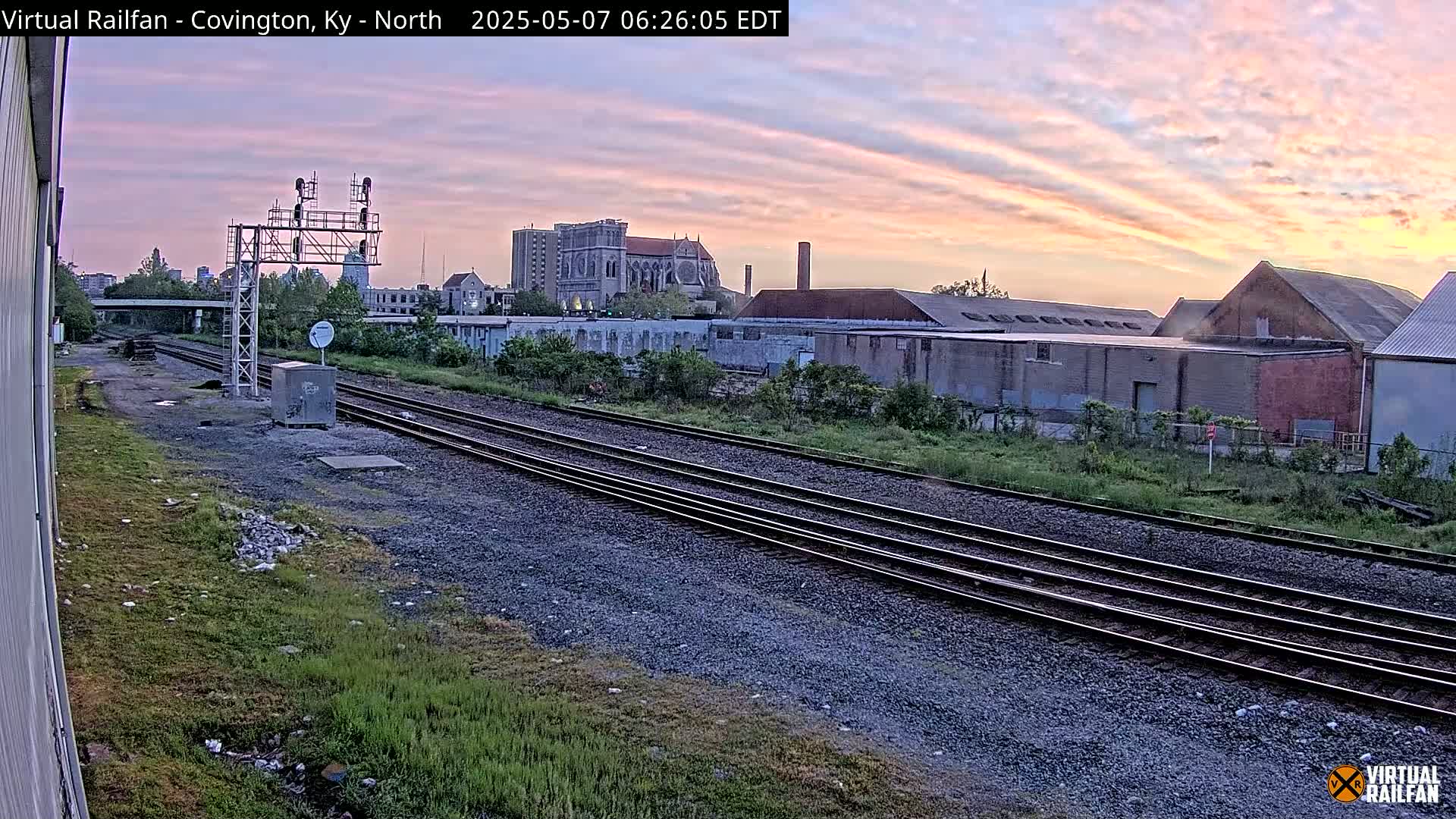 A view of railroad tracks at sunrise, with industrial buildings and a church visible in the background under a pale pink and orange sky.