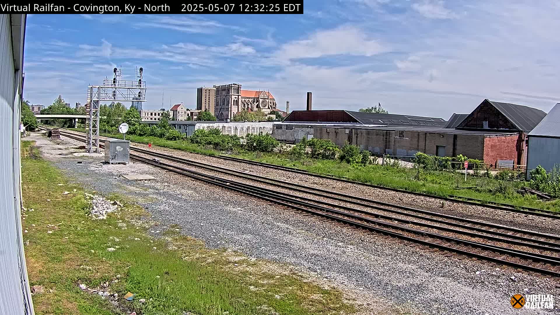 Under a partly cloudy sky, railroad tracks stretch toward a cityscape featuring industrial buildings and a large church.