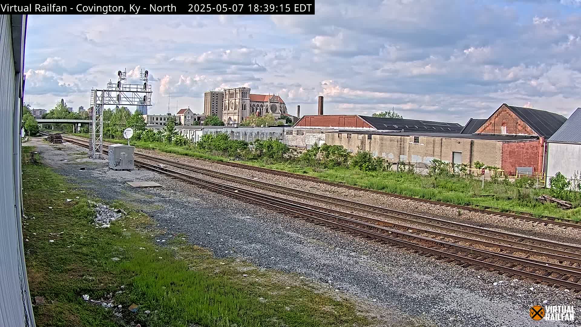 Railroad tracks stretch toward a cityscape under a partly cloudy sky, with industrial buildings and a large church visible in the distance.