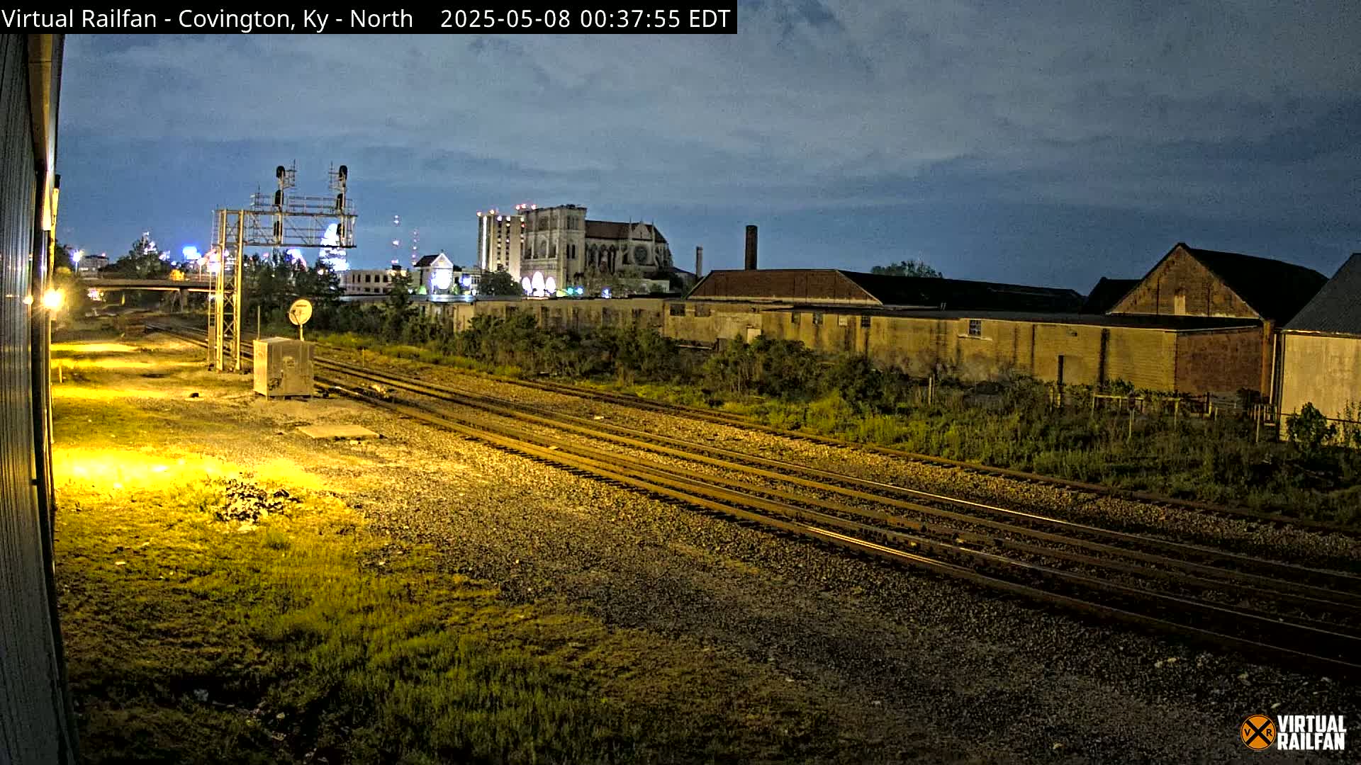 Under a twilight sky, multiple train tracks extend into the distance past industrial buildings and vegetation at night.