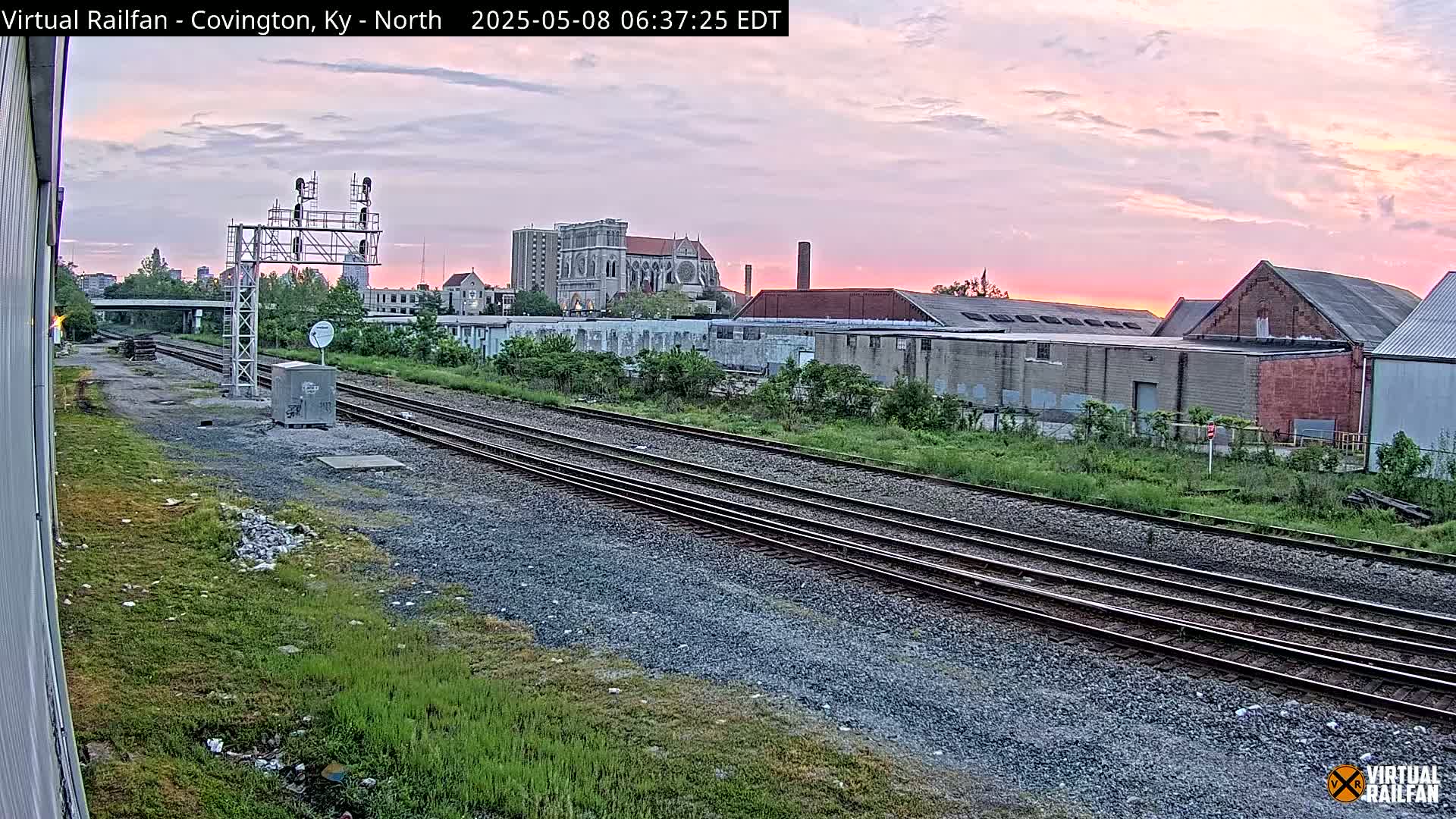 Multiple sets of train tracks stretch into the distance beneath a pinkish sunrise, past industrial buildings and a church.