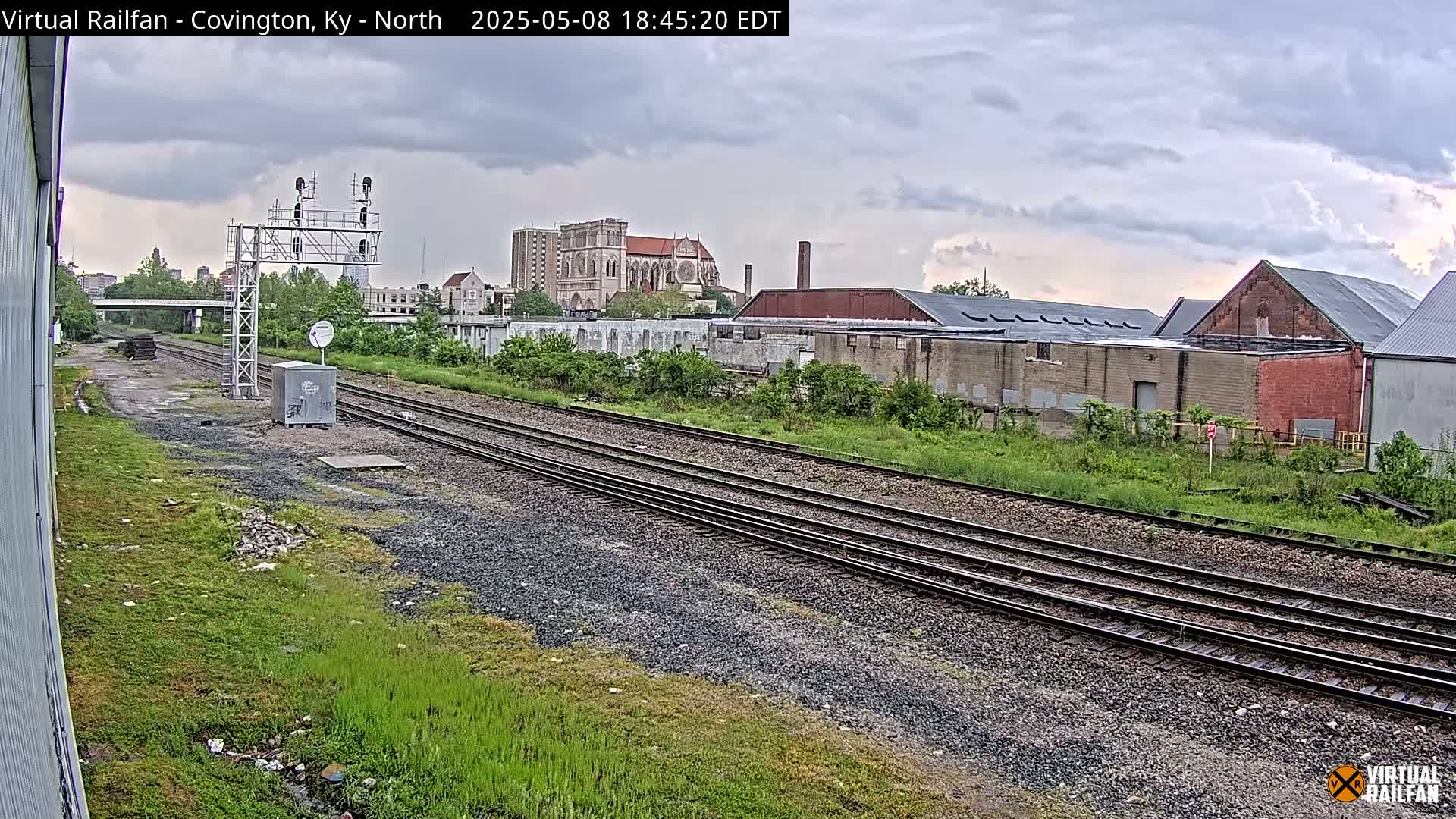 Overcast skies loom over railroad tracks that run alongside overgrown vegetation and industrial buildings, with a church visible in the distance.