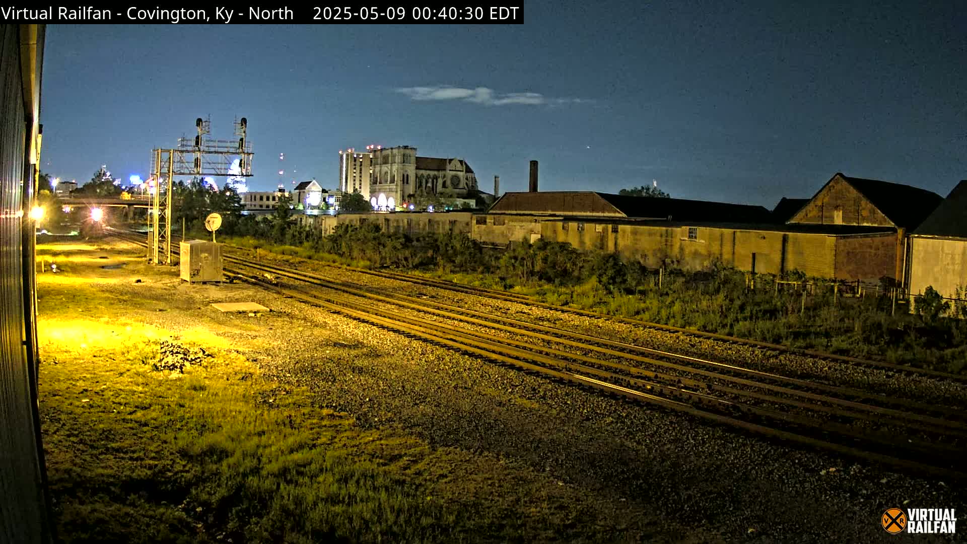 Under a night sky, multiple train tracks run through a grassy area toward a cityscape featuring buildings, including a large cathedral, under low light conditions.