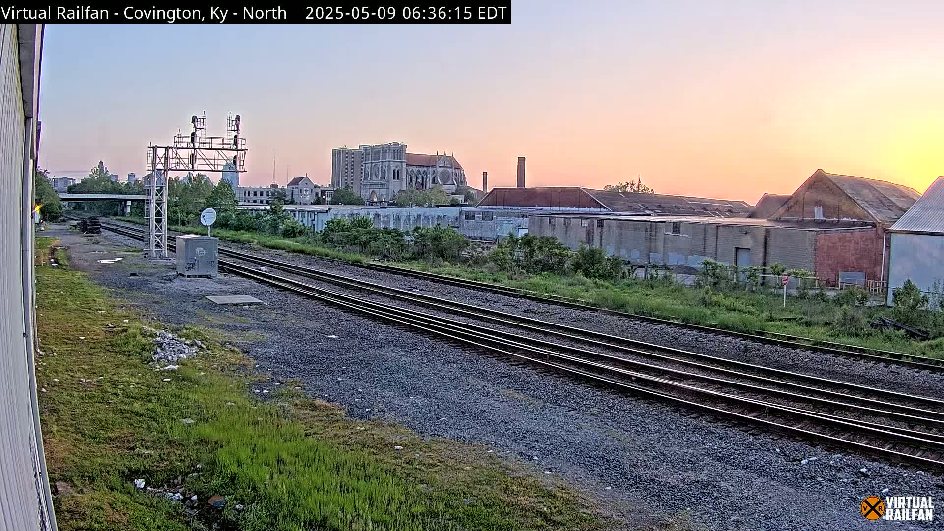Multiple train tracks stretch into the distance beneath a sunrise sky, past industrial buildings and a church.
