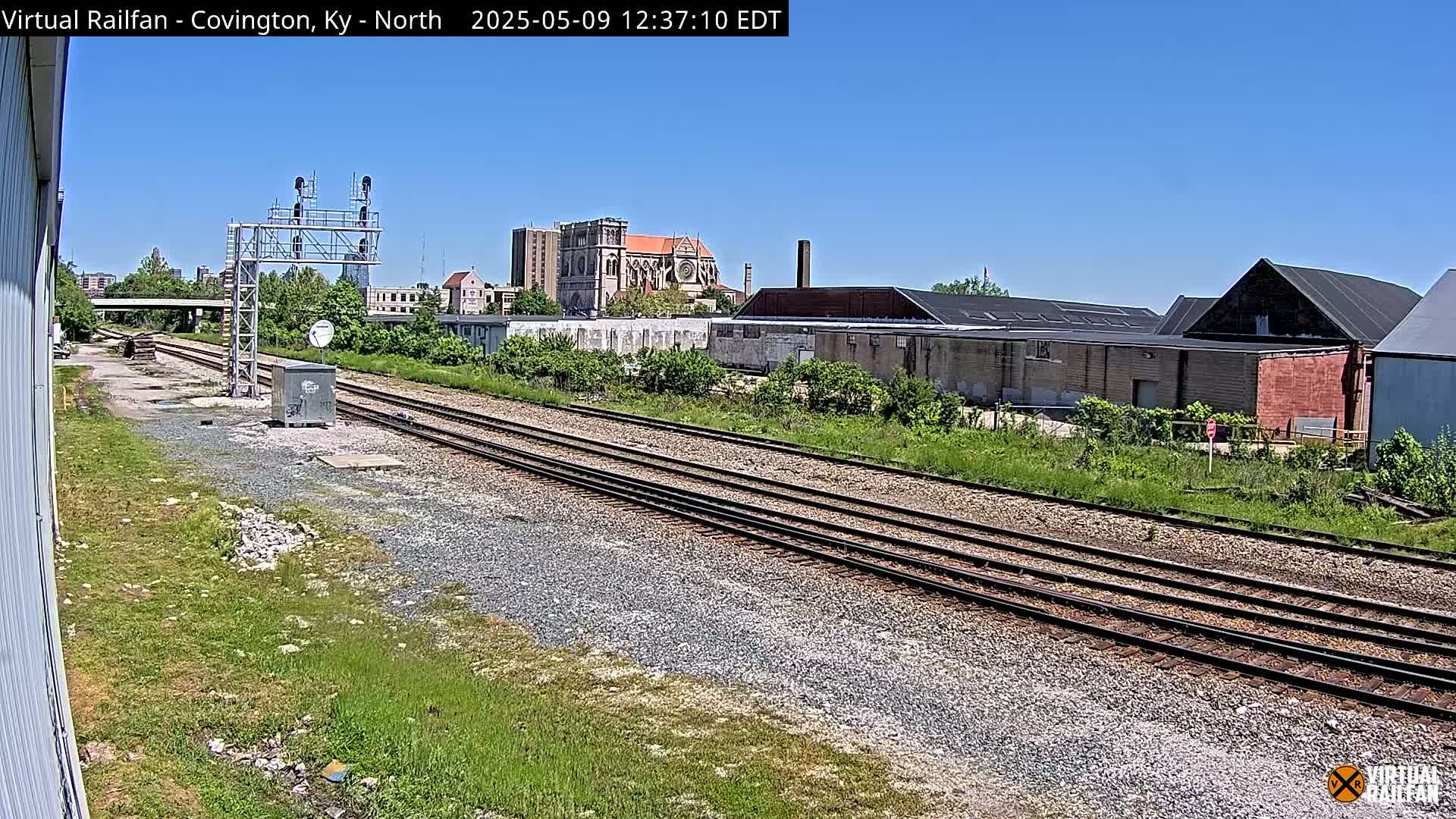 Under a clear blue sky, multiple train tracks stretch into the distance, passing industrial buildings and a church in the background.
