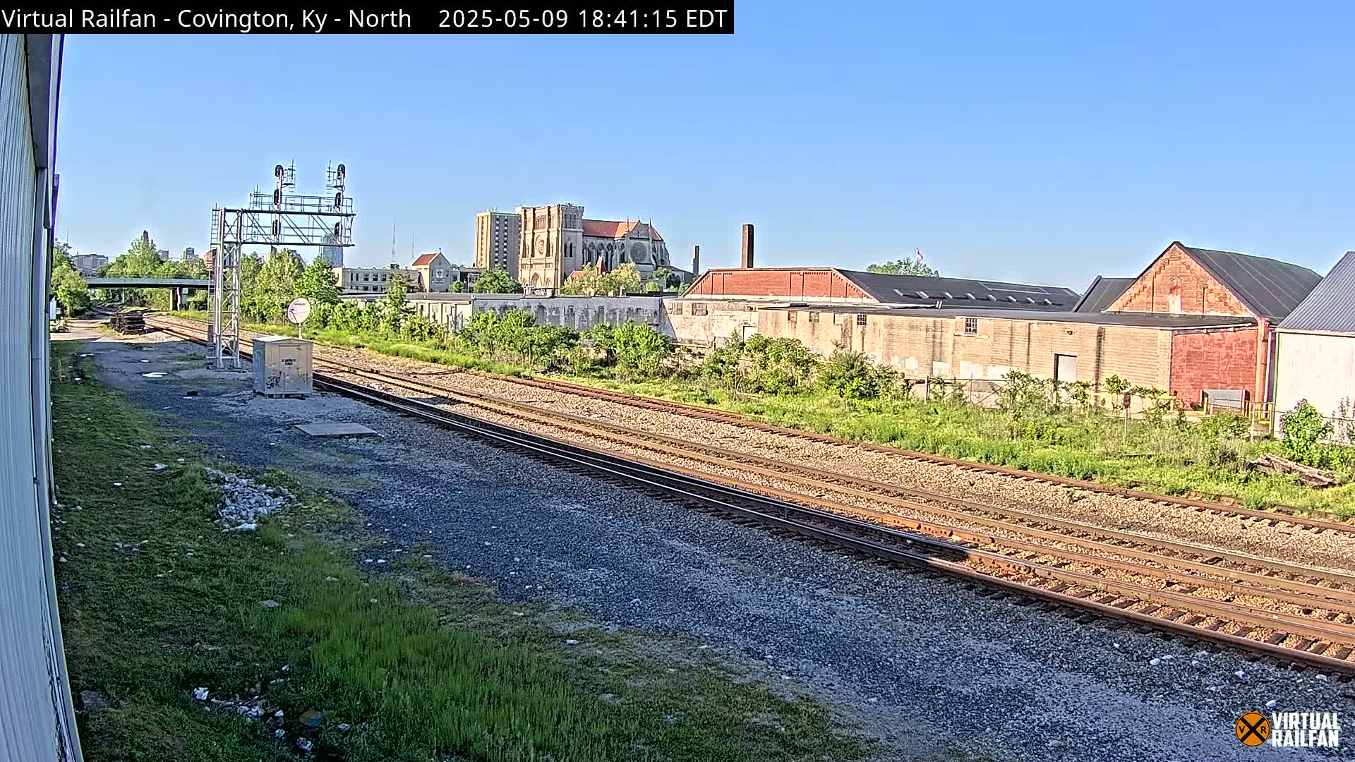 Multiple train tracks run through a grassy area past industrial buildings and a cathedral under a clear, sunny sky.