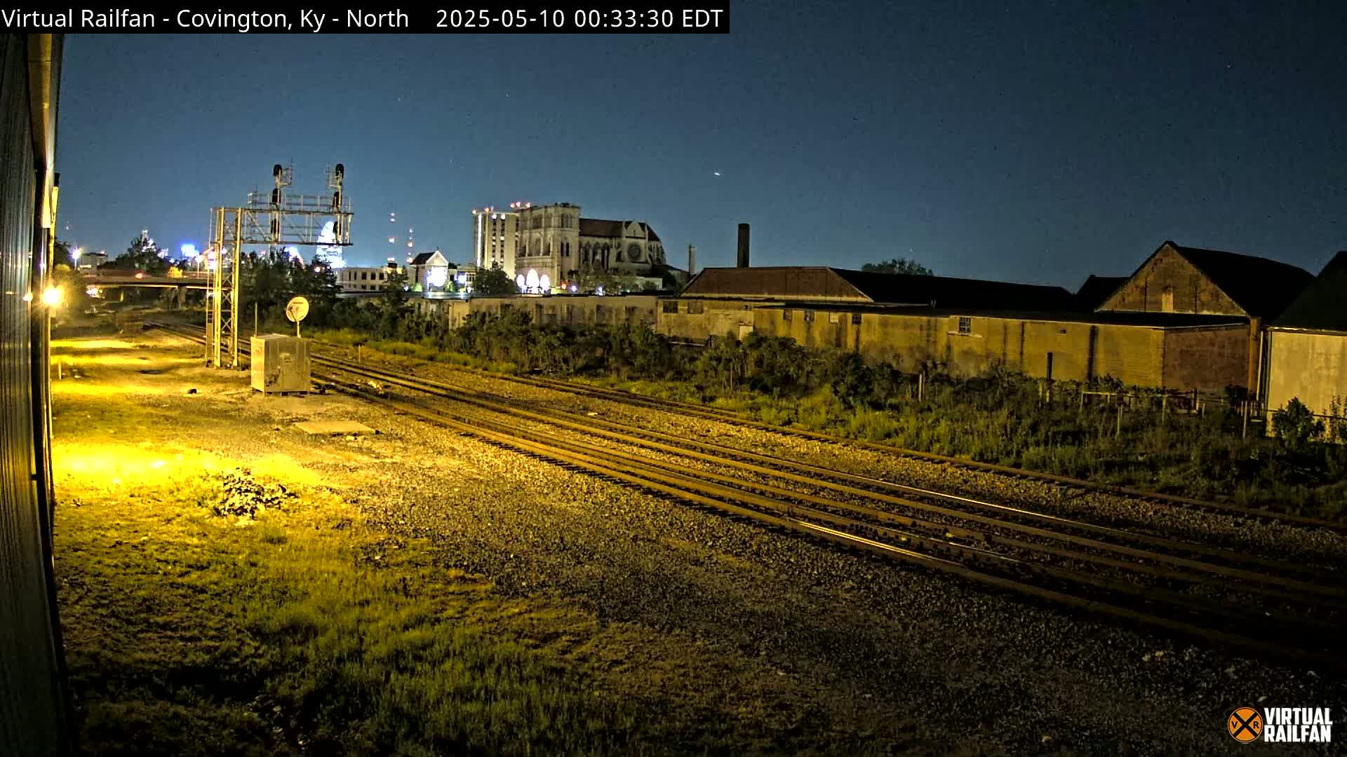 At night, under a clear sky, multiple sets of train tracks run past several low buildings and a large cathedral in the distance, illuminated by artificial light.