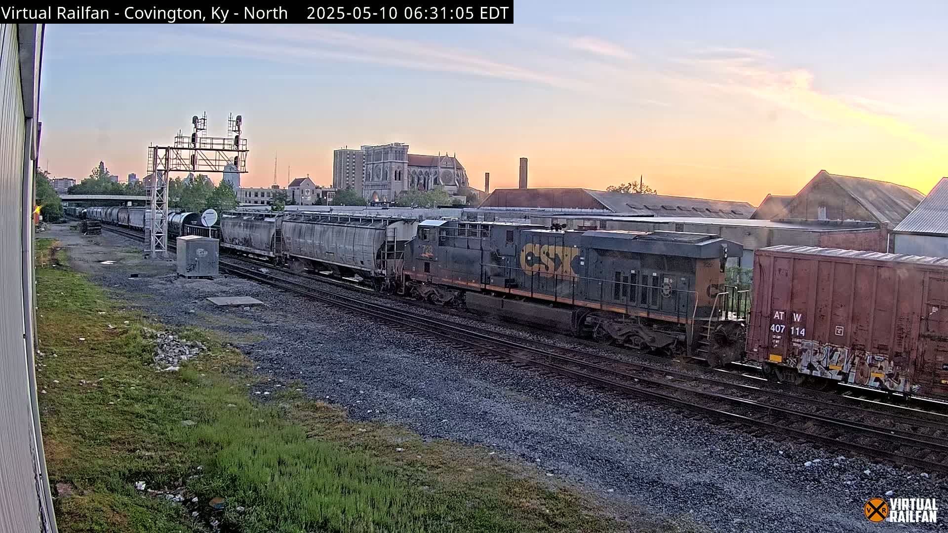 A freight train sits on railroad tracks at sunrise, with buildings and a church visible in the background.