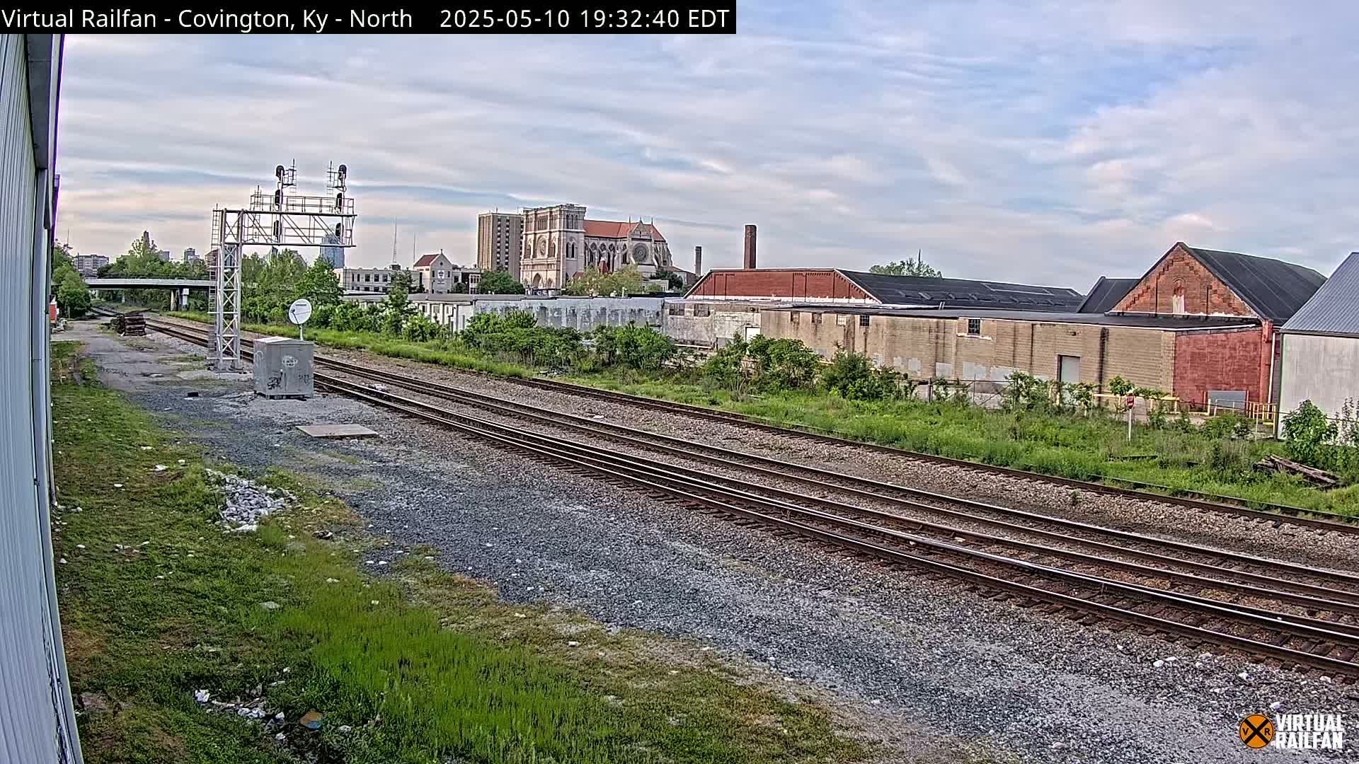 A view of railroad tracks on a partly cloudy day stretches toward a cityscape featuring industrial buildings and a large cathedral.