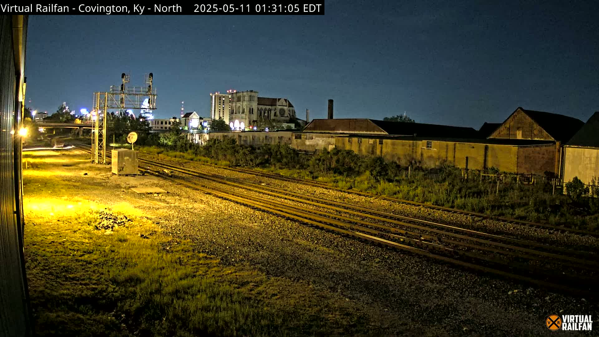 At night, under a clear sky, multiple train tracks extend into the distance past industrial buildings and a large cathedral.