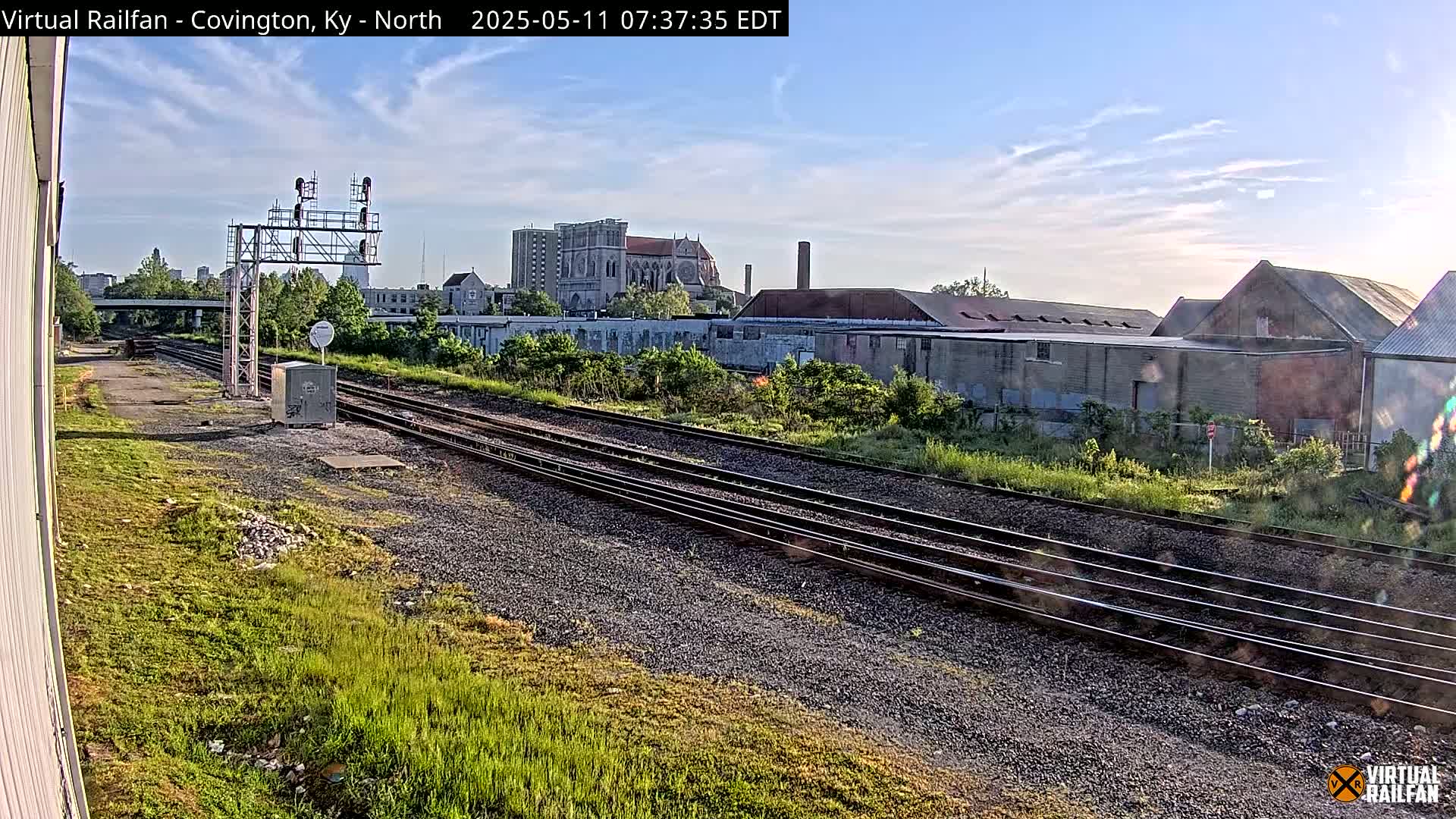 Multiple sets of train tracks run through a grassy area toward buildings and a church under a partly cloudy sky.