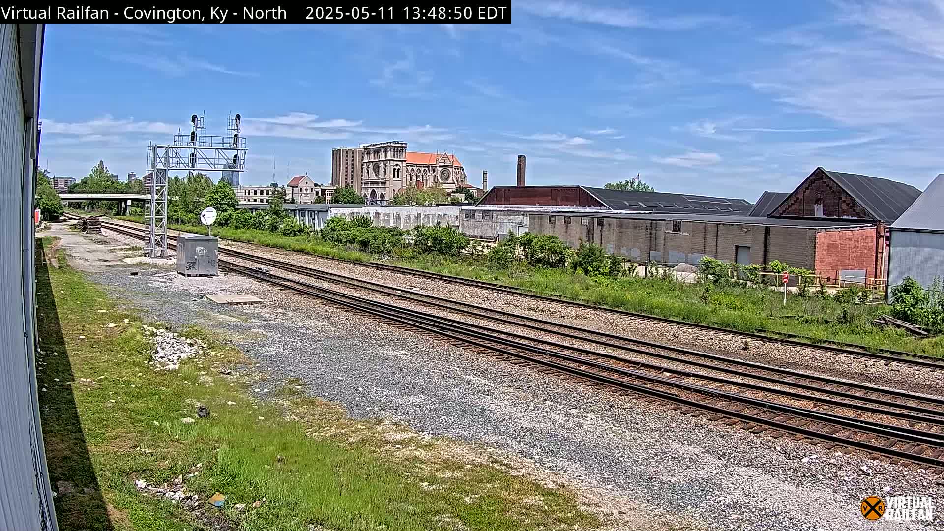 A sunny day reveals multiple train tracks extending into the distance, passing industrial buildings and a church, with lush green vegetation alongside.