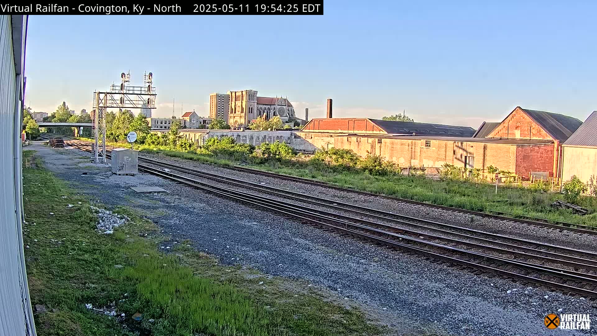 Under a clear sky, railroad tracks stretch towards a collection of brick buildings and a large cathedral in the distance.