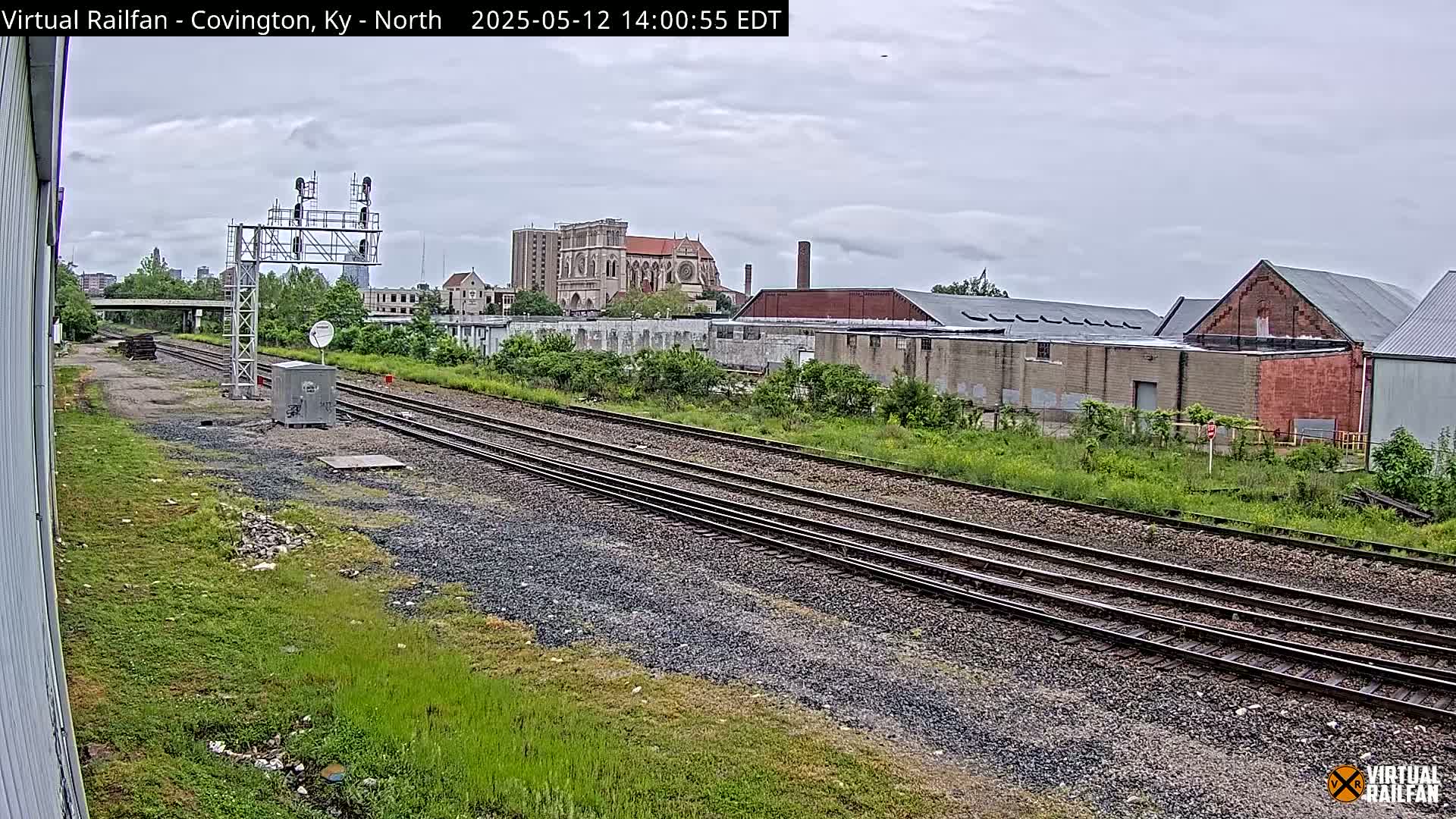 Multiple sets of train tracks stretch into the distance past overgrown vegetation and industrial buildings under an overcast sky.
