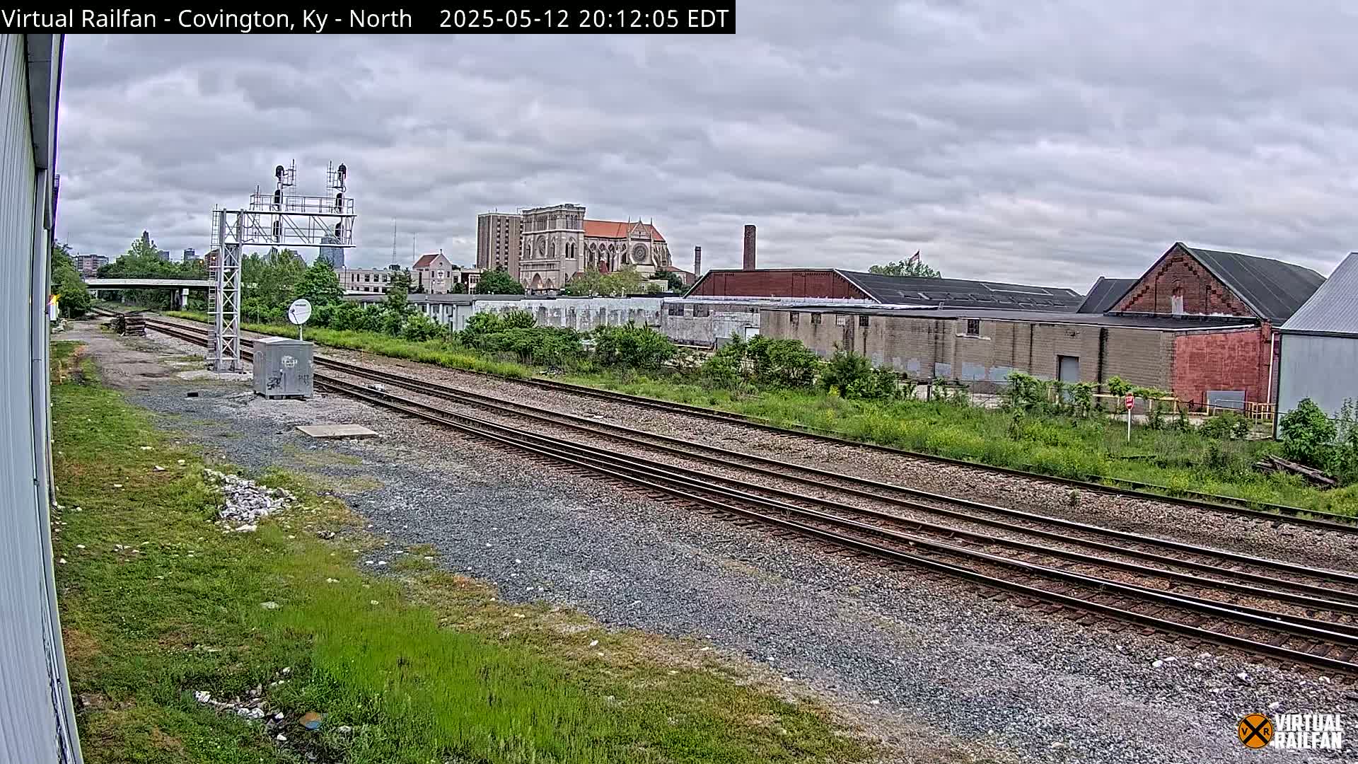 A view of railroad tracks under an overcast sky, with industrial buildings and a large church visible in the background.