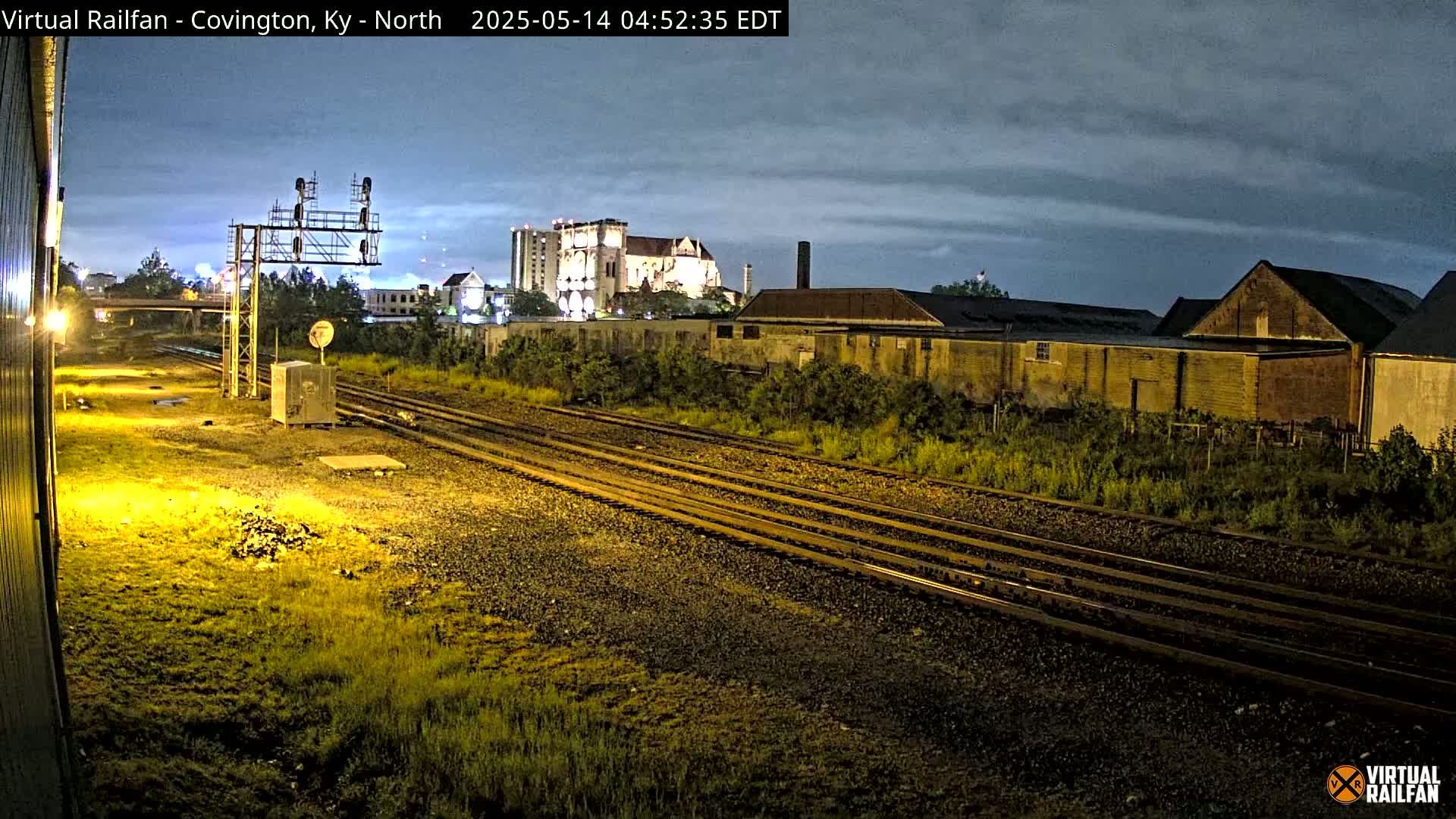 Under a partly cloudy night sky, several sets of train tracks run through a grassy area toward buildings and a large structure in the distance.