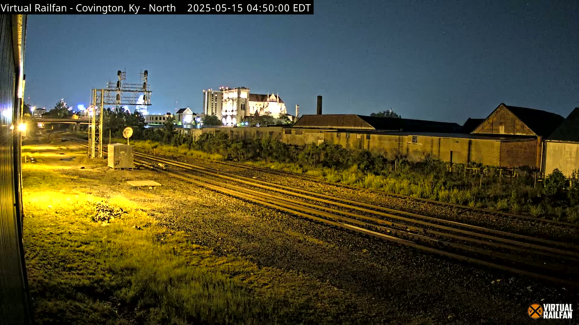 At night, multiple sets of train tracks extend from the foreground into the distance past buildings and overgrown vegetation under a clear sky.