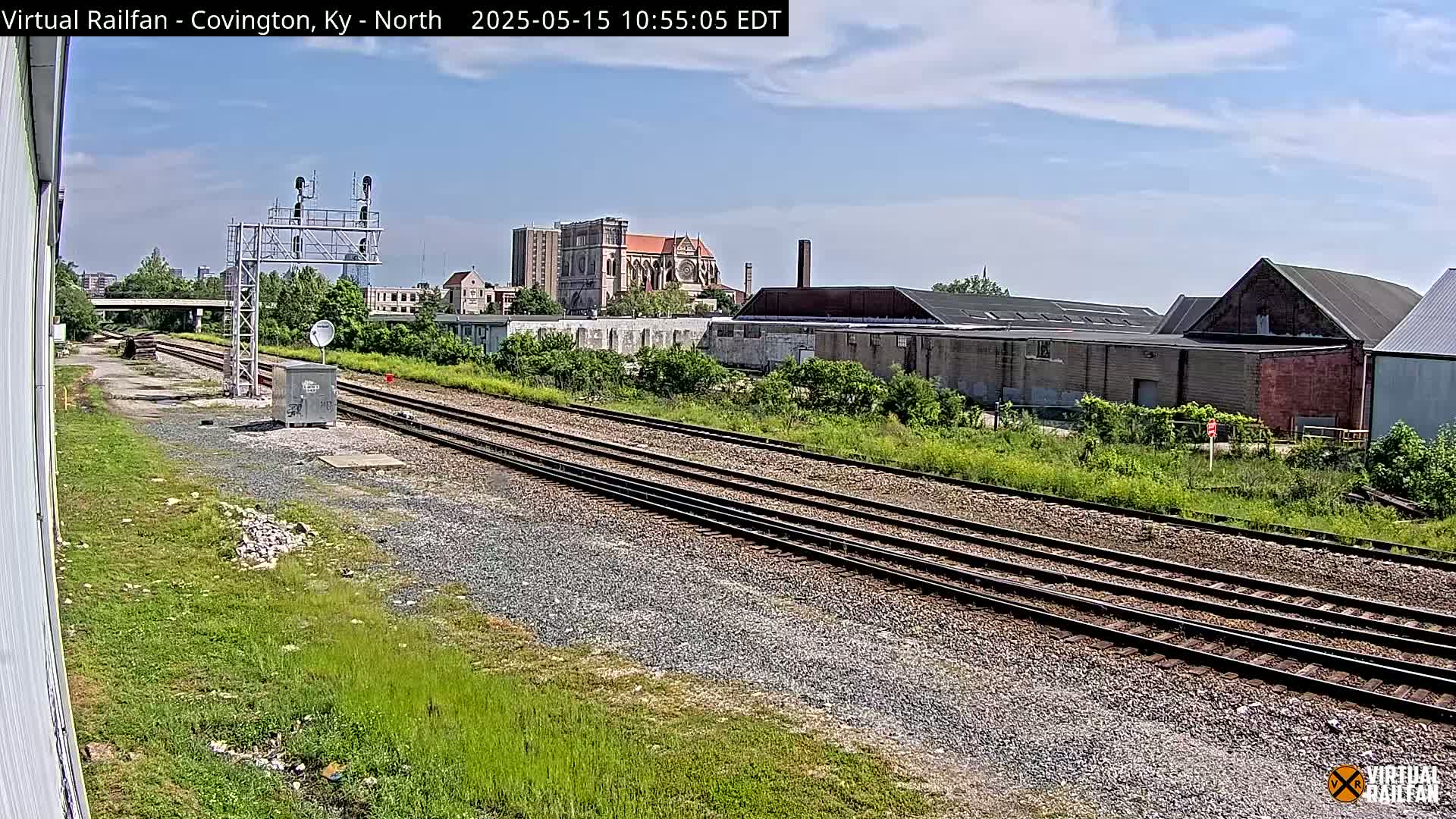 A sunny day view of train tracks extending into the distance, past industrial buildings and a church, with lush green vegetation alongside.