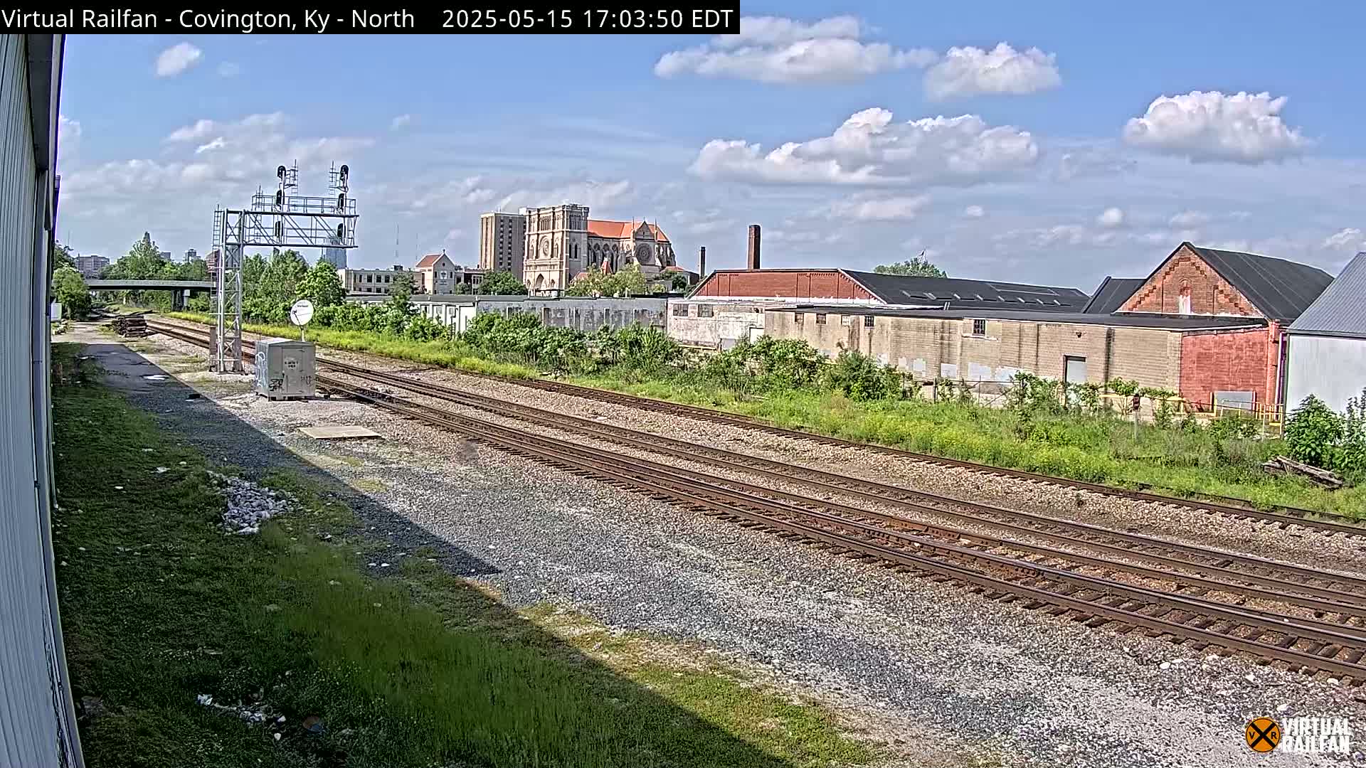 A sunny day reveals a railway track leading toward industrial buildings and a large cathedral under a partly cloudy sky.
