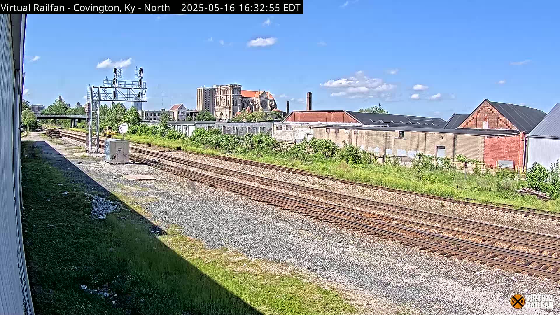 A view of railroad tracks on a sunny day, with industrial buildings and a church visible in the background.