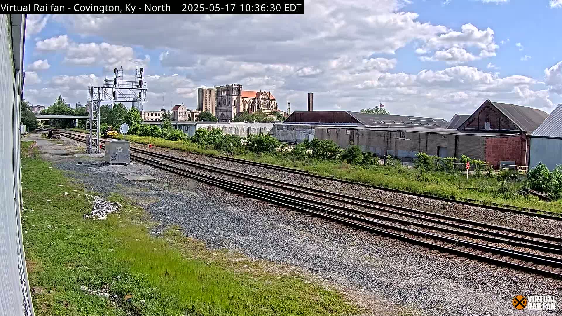 Under a partly cloudy sky, railroad tracks extend into the distance past industrial buildings and a large church.