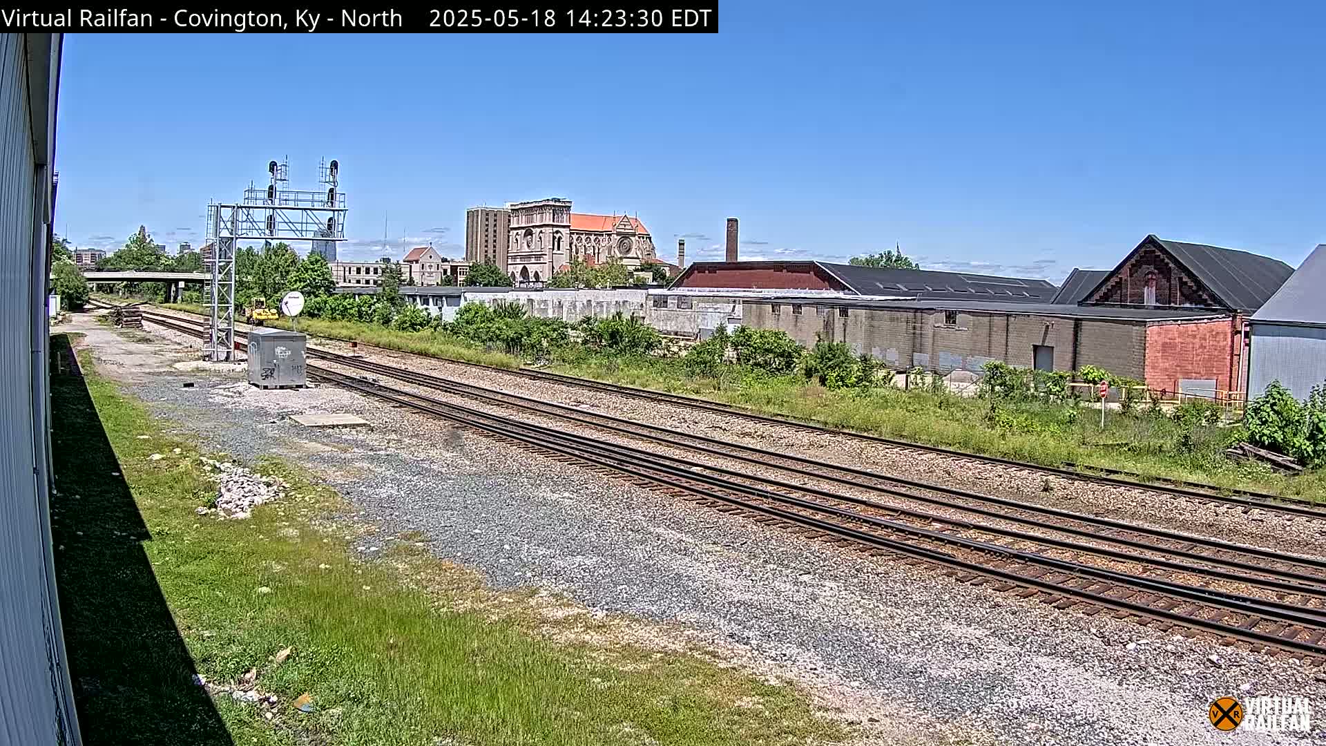 Under a clear blue sky, a view of railroad tracks stretches into the distance, passing by industrial buildings and a church in the background.