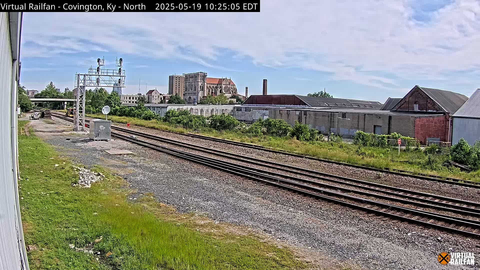 Multiple sets of train tracks run alongside overgrown vegetation, past industrial buildings and a large church under a partly cloudy sky.