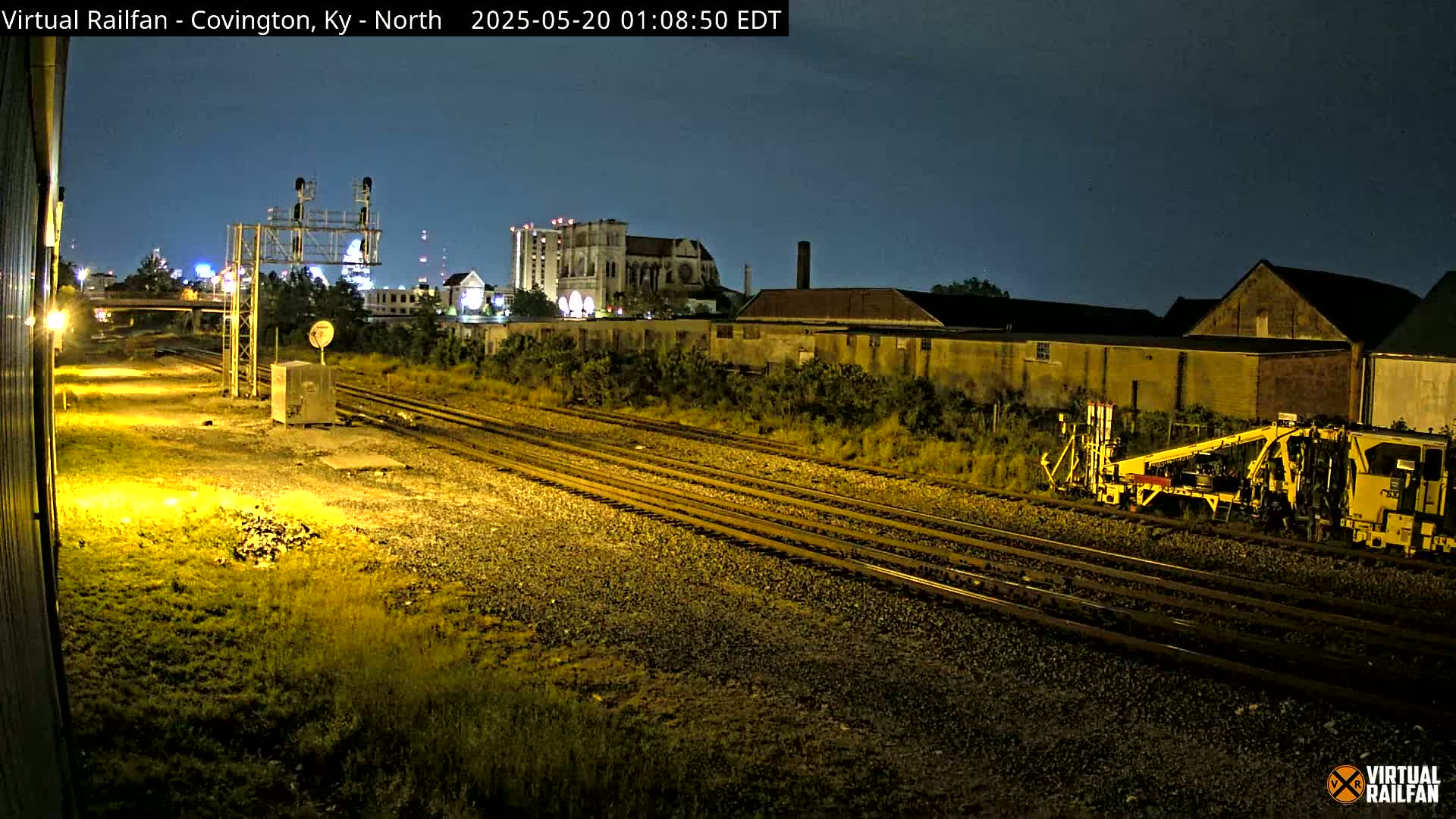 At night, under a clear dark sky, a yellow railway maintenance vehicle sits beside several train tracks that run past buildings and overgrown vegetation.
