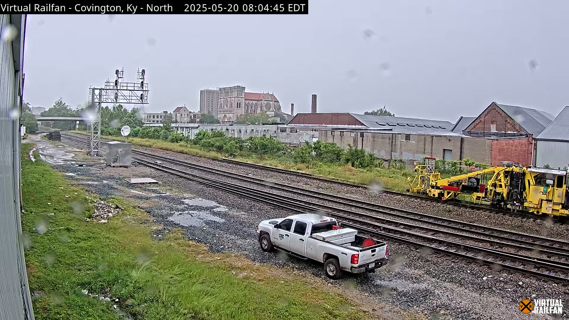 A rain-soaked railway scene shows a white pickup truck parked near a yellow rail maintenance vehicle on multiple train tracks, with industrial buildings and a church in the background.