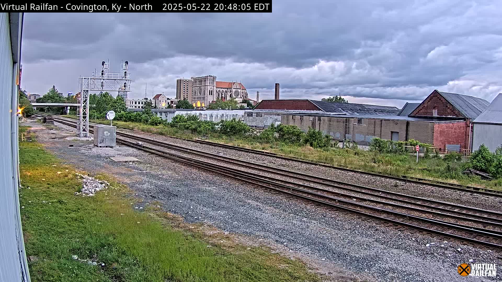 Multiple train tracks extend into the distance under a cloudy sky past industrial buildings and a large cathedral.