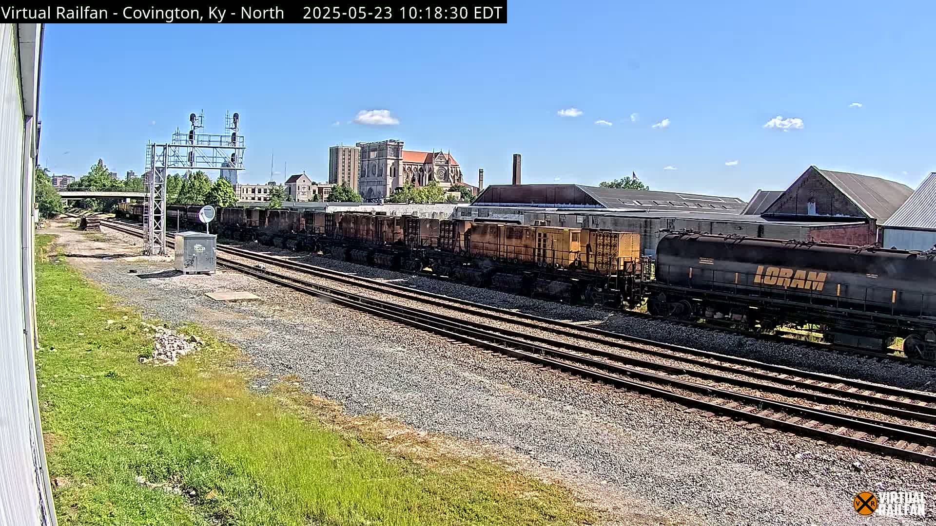 A long freight train sits on a railway track under a clear, sunny sky, with buildings and a church visible in the background.