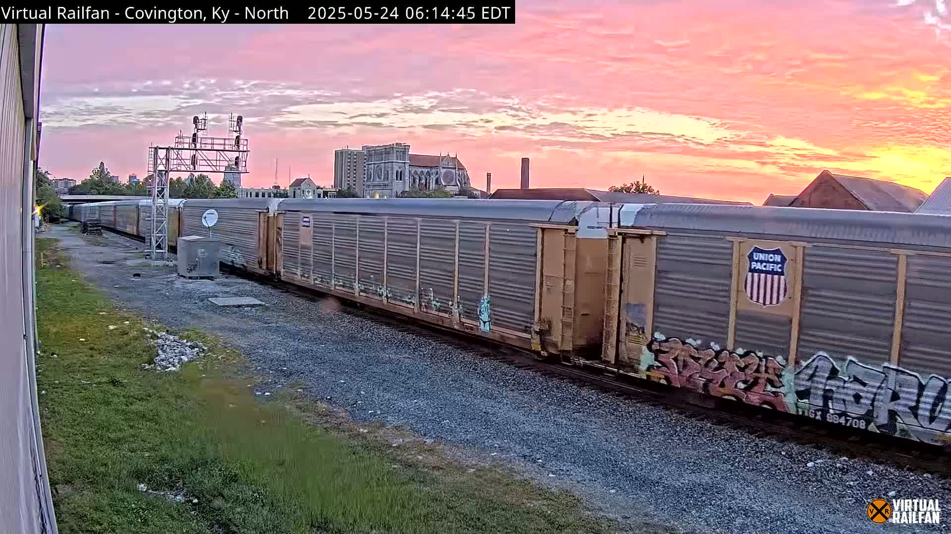 A long freight train sits on the tracks during a sunrise with a pink and orange sky.