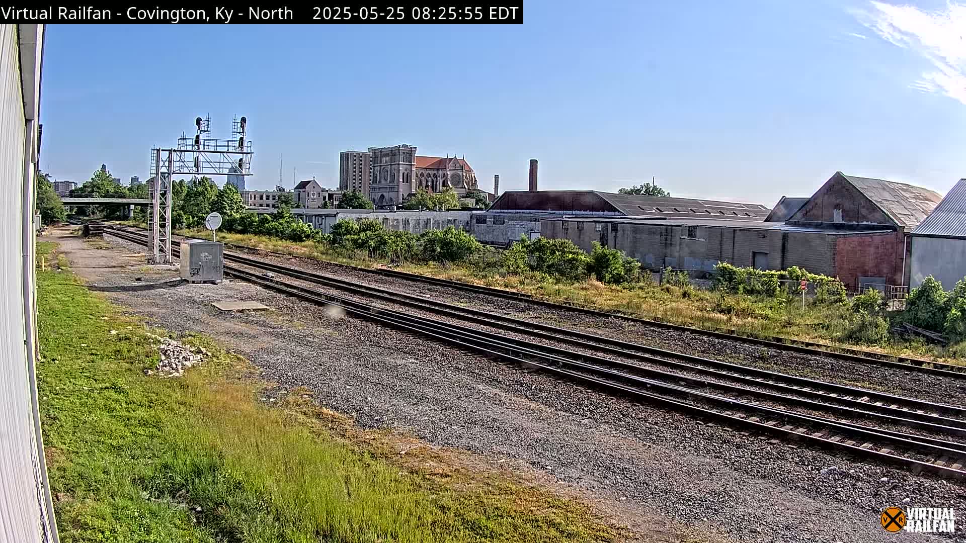 Multiple train tracks run alongside industrial buildings and overgrown vegetation under a clear blue sky.