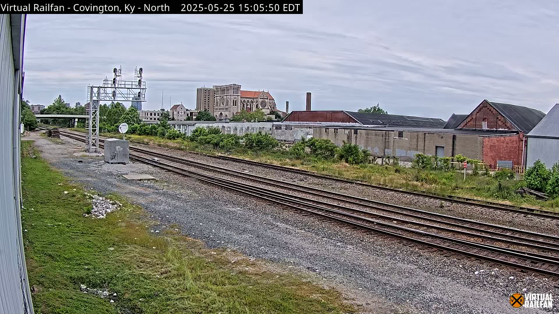 Under an overcast sky, a view of railroad tracks stretches towards a cityscape featuring industrial buildings and a large church in the background.