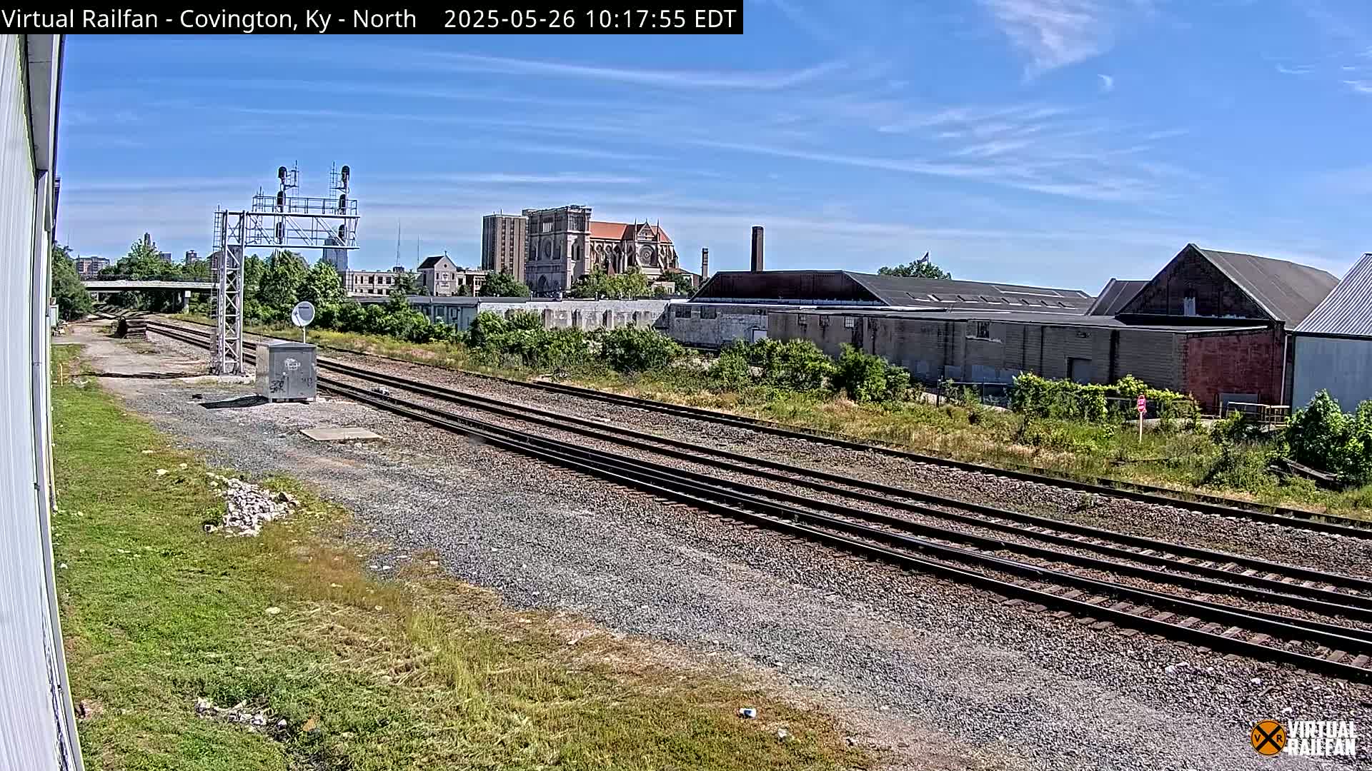 A sunny day reveals multiple train tracks stretching into the distance, past industrial buildings and a large cathedral under a mostly clear blue sky.
